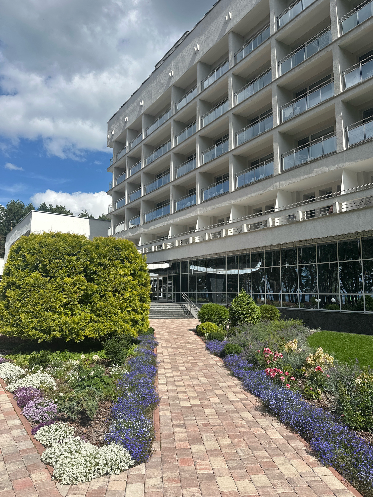 The image shows the exterior of a multi-story hotel building with a modern, white facade. The building features numerous balconies with glass railings and is surrounded by a well-maintained garden and a paved walkway. In the foreground, a vibrant green bush is prominent, and flowerbeds with a variety of colorful flowers, including purple, white, and pink blooms, line the path. The sky above is partly cloudy with blue sky visible. The scene suggests a resort or hotel located in a natural setting, possibly in the city of Мядзельскі сельскі Савет, Belarus, given the context. There are no people visible in the image.