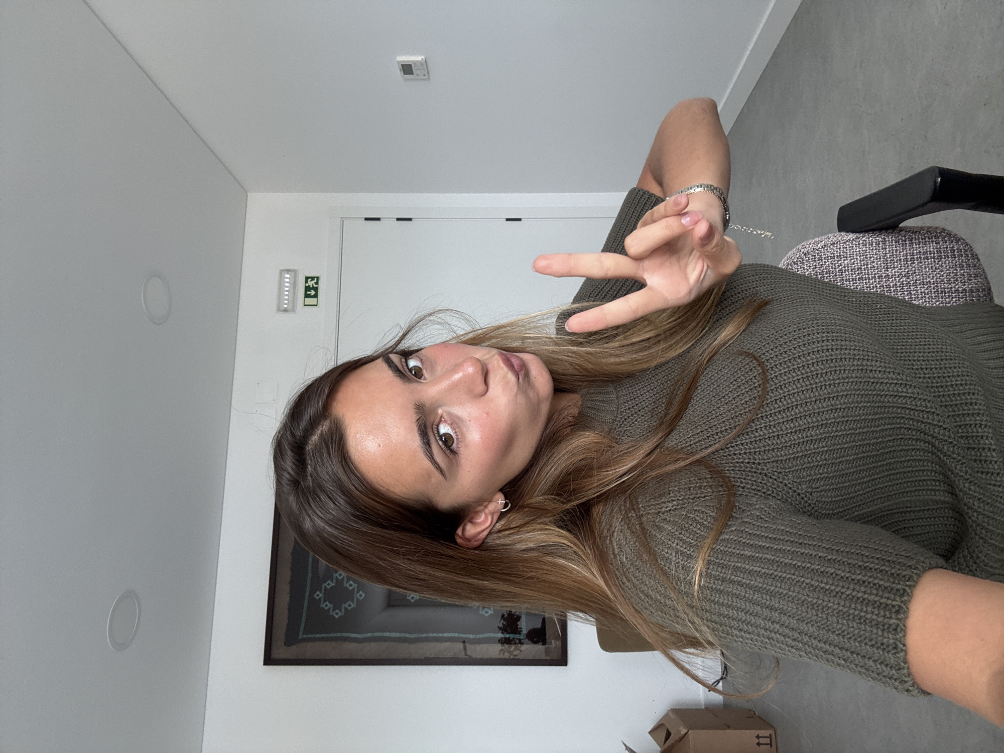 A young woman is captured in a selfie, with her head tilted to the side and a slight pucker on her lips, looking directly at the camera. She holds up two fingers in a peace sign with her left hand, which displays a silver-colored bracelet. Her long, brown hair with lighter highlights frames her face and falls over her shoulders. She is wearing a textured, dark green knit sweater. A small, silver-toned cross earring is visible in her right ear.
The setting appears to be a modern indoor space, possibly an office or a bright room in Torres Vedras, Portugal. The walls and ceiling are white, providing a clean backdrop. In the background, a white door is visible, above which a green emergency exit sign with an arrow pointing left and a human figure can be seen. A white square device, possibly a smoke detector, is mounted on the ceiling, along with a circular, recessed light fixture. A framed artwork featuring a dark geometric pattern hangs on the wall behind her left shoulder. Part of a grey office chair is visible to her right, and a brown cardboard box with faint markings, including "II" in a white square, is present in the lower right corner. The flooring appears to be a grey, possibly concrete or tiled surface. The lighting is bright and even, consistent with an interior space during the day. The text "happe" is indistinctly visible on the silver bracelet.