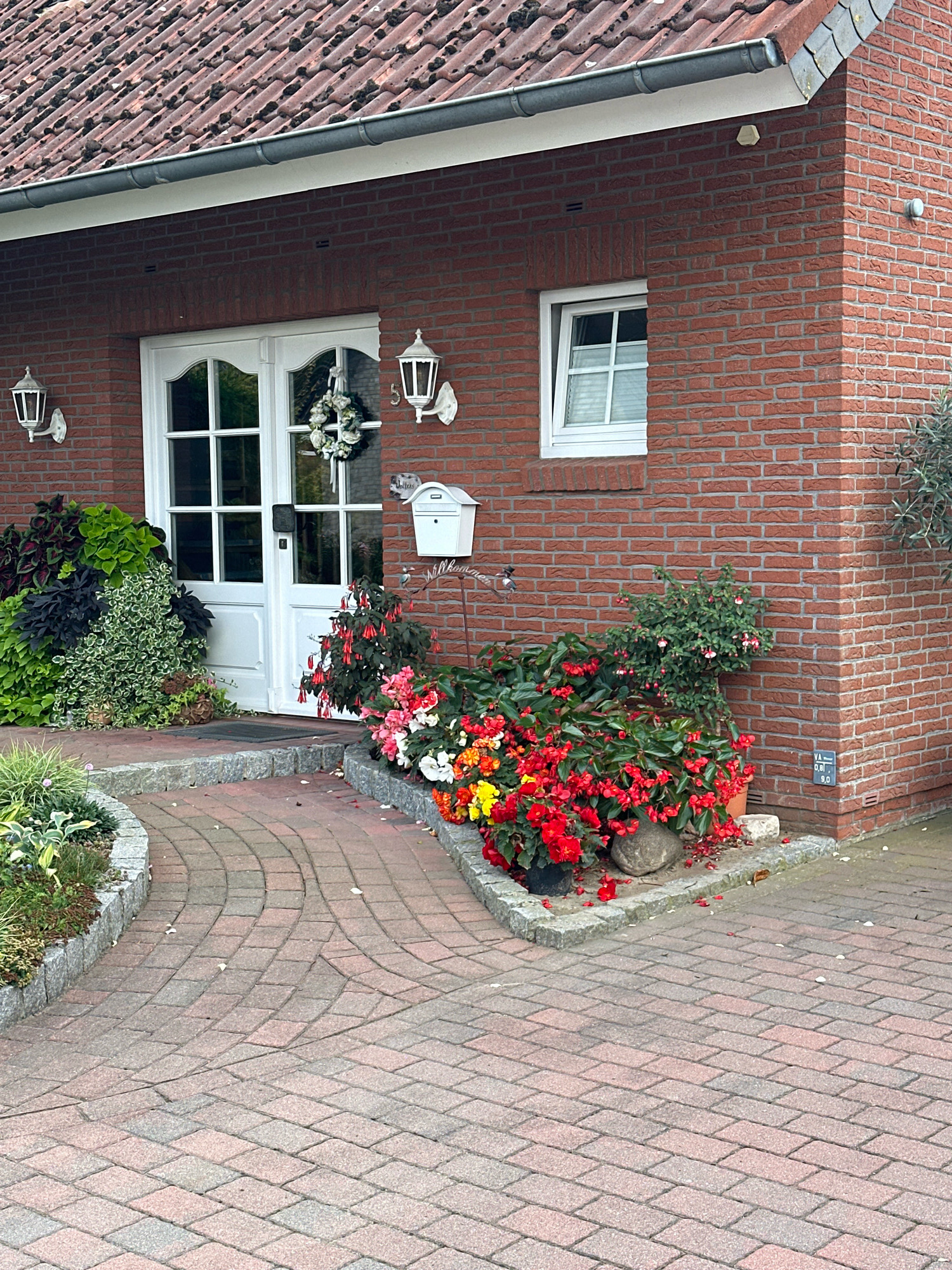 The image depicts the front facade of a two-story brick house in Samtgemeinde Thedinghausen, Germany. The house has a red tile roof with visible moss. A white front door with glass panes is centered on the facade. Flanking the door are two white outdoor lanterns. To the left of the door, a dense arrangement of green and dark purple foliage is planted. To the right of the door, a white mailbox is mounted on the brick wall. Below the mailbox and to the right of the door, a cluster of red and pink flowers, likely begonias and fuchsias, are in bloom. The walkway leading to the door is paved with red and gray interlocking bricks, forming a curved path. In the foreground, more of the brick pathway is visible, with some scattered leaves. The overall scene presents a well-maintained entrance to a residential property.