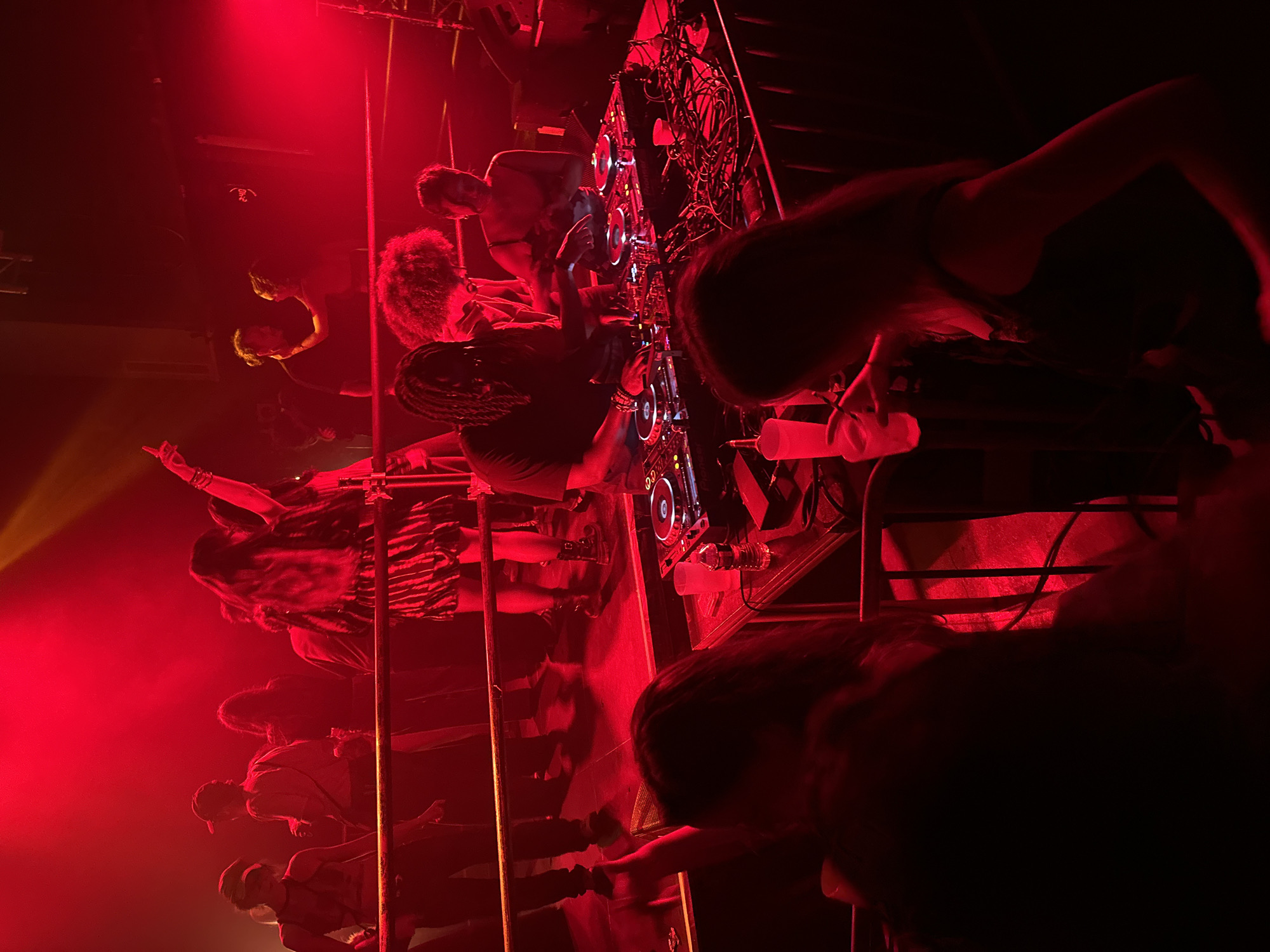 A low-angle shot depicts multiple people seemingly on an elevated stage, likely performing in a nightclub, possibly in Paris, France. The scene is lit with a dominant red light, which obscures details. Several individuals are visible from the chest up, possibly DJs or performers manipulating equipment such as turntables and wires. The crowd is below, partially obscured by the darkness. Metal scaffolding and vertical bars are also noticeable. The lighting creates a sense of dynamism and intensity in the scene.