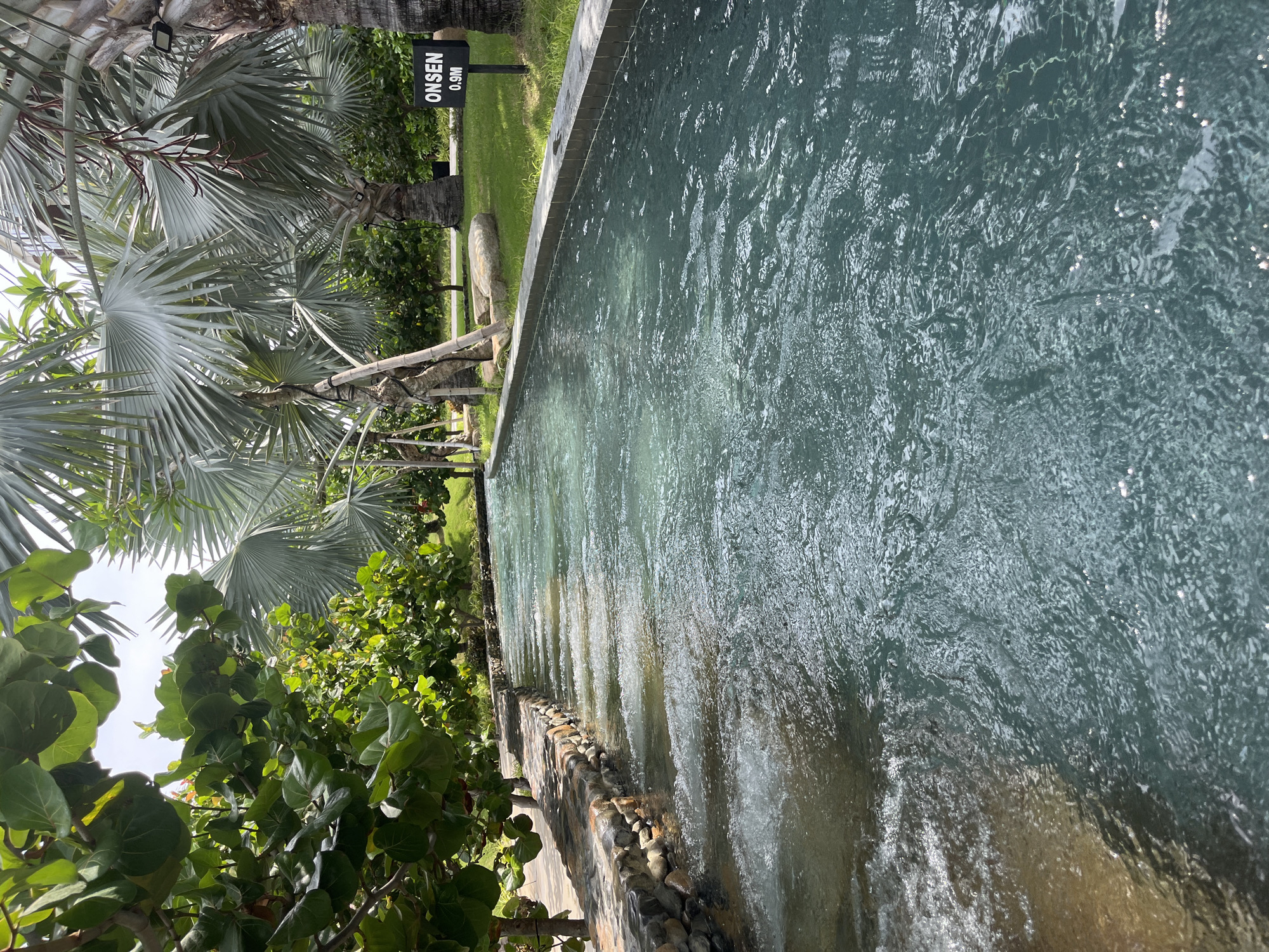 The image depicts a man-made water channel flowing through a tropical landscape. The channel is lined with stone and concrete, with water flowing gently, creating ripples and small waves. On the left side of the channel, there is a stone wall with lush green foliage overhanging. On the right side, the channel is bordered by a well-manicured lawn, with palm trees and other tropical vegetation visible in the background. A black sign with white lettering reads "ONSEN 0.9M", indicating a depth of 0.9 meters and that this is likely an onsen, or hot spring. The sky is overcast, suggesting it might be a humid or partly cloudy day. The overall scene conveys a sense of tranquility and relaxation, typical of a resort or spa environment in a warm climate. The location is identified as Cam Lam, Vietnam. There are no people visible in the image.