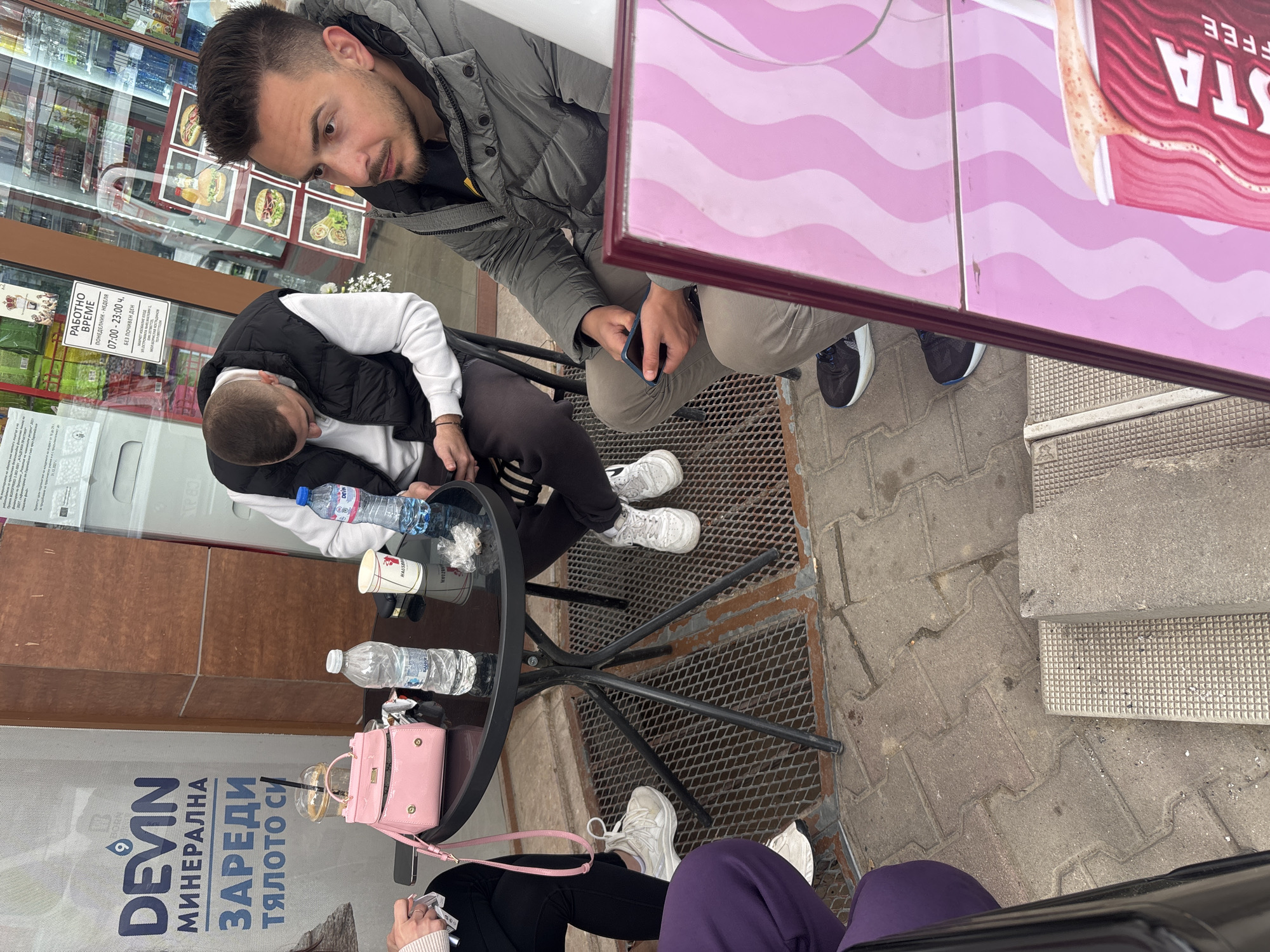 Here is a concise description of the image:
The image captures an outdoor scene in Plovdiv, Bulgaria, likely a casual gathering at a Costa Coffee establishment. Three people are visible around a small black table. One man is looking into the lens, wearing a grey jacket and khaki pants. Another man wearing a white shirt and black vest is seated at the table looking down. A third person, only partially visible, is sitting next to the pink purse, wearing black pants and purple pants. Objects on the table include water bottles, a paper coffee cup, and a plastic cup. A pink purse and pack of cigarettes are placed close to the base of the table. The location is identifiable by Bulgarian writing on the shopfront, and Costa Coffee signage on the table, indicating the cafe's branding.