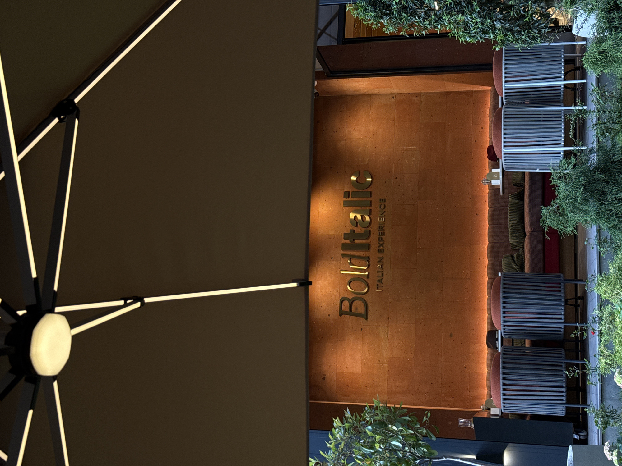 This image captures an inviting outdoor dining or lounge area, likely for a restaurant or cafe, at night or in the late evening, suggested by the warm ambient lighting. The scene, located in Yerevan, Armenia, features a sophisticated design.
On the left side of the frame, a large, dark-colored patio umbrella extends, its internal support ribs illuminated with prominent white LED strips that provide a soft, appealing glow. The structure of the umbrella suggests a modern design.
To the right, a building facade made of a warm, reddish-brown textured material serves as the background. Prominently displayed on this facade is the name "Bolditalic" in elegant, golden, three-dimensional lettering. Below the main name, the tagline "ITALIAN EXPERIENCE" is written in smaller, golden capital letters, indicating the type of cuisine or establishment.
Below the illuminated sign, an outdoor seating area is arranged. It features comfortable-looking sofas with patterned upholstery and several contemporary chairs with slatted backs and reddish cushions. Small tables are placed between the seating arrangements, with one table visible holding a glass decanter. Lush green plants and foliage are strategically placed around the perimeter and within the seating area, adding a touch of nature and enhancing the relaxed atmosphere. The combination of warm lighting, comfortable seating, and greenery creates an inviting and upscale ambiance for patrons. No people are visible in the scene.