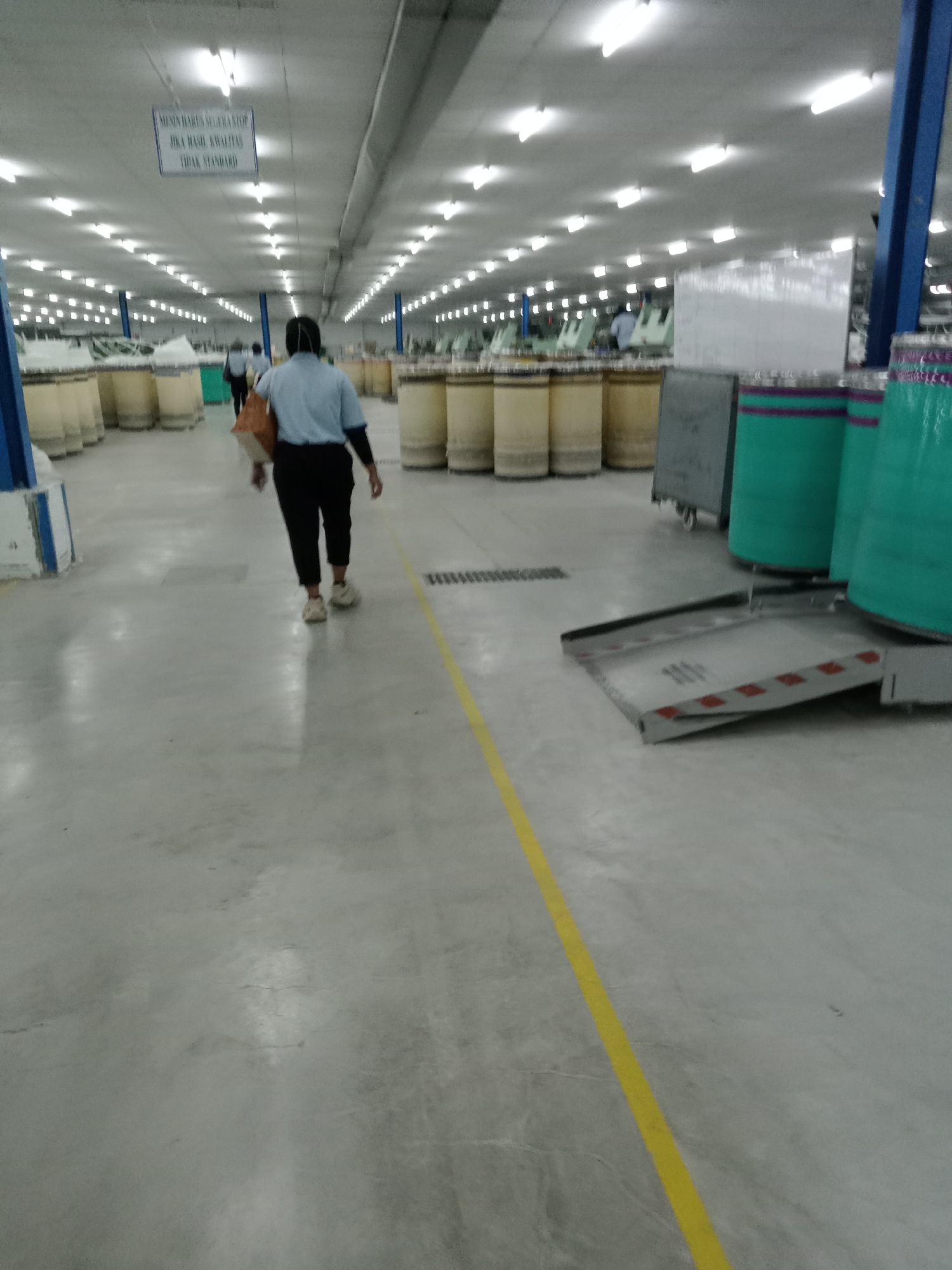 This image depicts the interior of a large industrial facility, likely a textile or yarn production factory, located in Nglundo, Indonesia. The scene is dominated by a wide, well-lit aisle with a light gray concrete floor, marked by a bright yellow line running down its center.
In the foreground, a person is seen from behind, walking away from the camera. They are dressed in a light blue short-sleeved shirt, dark trousers, and light-colored sneakers, carrying a light brown bag over their shoulder. Another person is partially visible further down the aisle, also appearing to walk.
On both sides of the aisle, rows of numerous large, cylindrical bobbins or spools, mostly tan or yellowish, are neatly arranged, suggesting the presence of yarn or thread. On the right side, closer to the viewer, are several taller cylindrical objects wrapped in teal plastic with purple bands, possibly larger spools or material containers. A gray industrial cart and a metal ramp with red and white striped markings, likely used for material handling, are also visible on the right.
The ceiling is brightly illuminated by numerous rectangular fluorescent lights, suggesting active operations regardless of external daylight. A white rectangular sign hangs from the ceiling in the mid-distance. The text on the sign, in Indonesian, reads: "MESIN HARUS SEGERA STOP JIKA HASIL KUALITAS TIDAK STANDARD," which translates to "MACHINE MUST STOP IMMEDIATELY IF QUALITY RESULTS ARE NOT STANDARD." This detail highlights the facility's emphasis on quality control and standard operating procedures within this manufacturing environment.