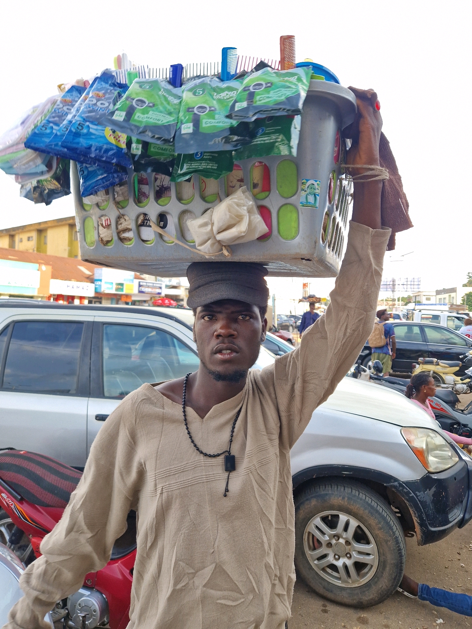 A street vendor carrying a large basket of merchandise on his head walks through a busy street. The vendor, a young man wearing a beige tunic and a dark cap, has his right arm raised, supporting the basket. The basket is filled with various items, including combs, what appear to be packaged sanitary products, and other small consumer goods. The scene takes place outdoors, with parked cars and motorcycles visible in the background, suggesting an urban environment. Several buildings are also in the distance, indicating a commercial area. The overall impression is that of a typical street market scene in a bustling city.