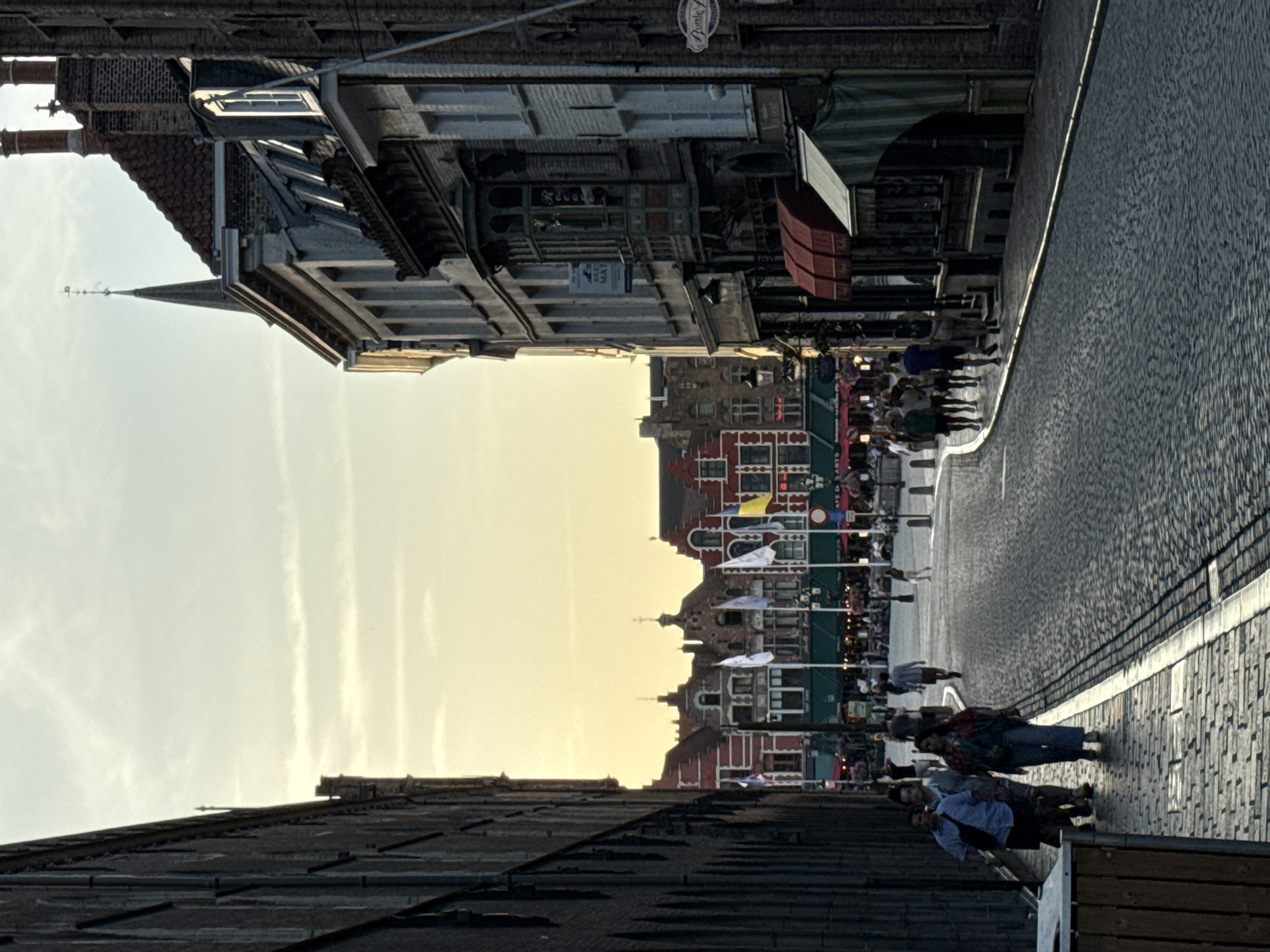 The image depicts a street scene in Bruges, Belgium, with buildings lining one side of a cobblestone street. The orientation of the image is such that the buildings appear on the left and the street on the right, with the sky and further buildings visible in the background.
The subjects include a group of people walking along the sidewalk and the street. The buildings showcase traditional European architecture with brick facades, ornate windows, and pitched roofs. Some buildings appear to be shops or cafes with awnings and displays. Flags are visible on poles in front of some buildings.
The time of day appears to be late afternoon or early evening, as suggested by the warm, soft light and the long shadows cast by the buildings. The sky is clear with some wispy clouds. The atmosphere seems relaxed and pleasant, with people enjoying an evening stroll.
Notable details include the texture of the cobblestone street and the detailed brickwork of the buildings. The overall impression is one of a charming, historic European city.
No visible text is clearly legible in the provided image.