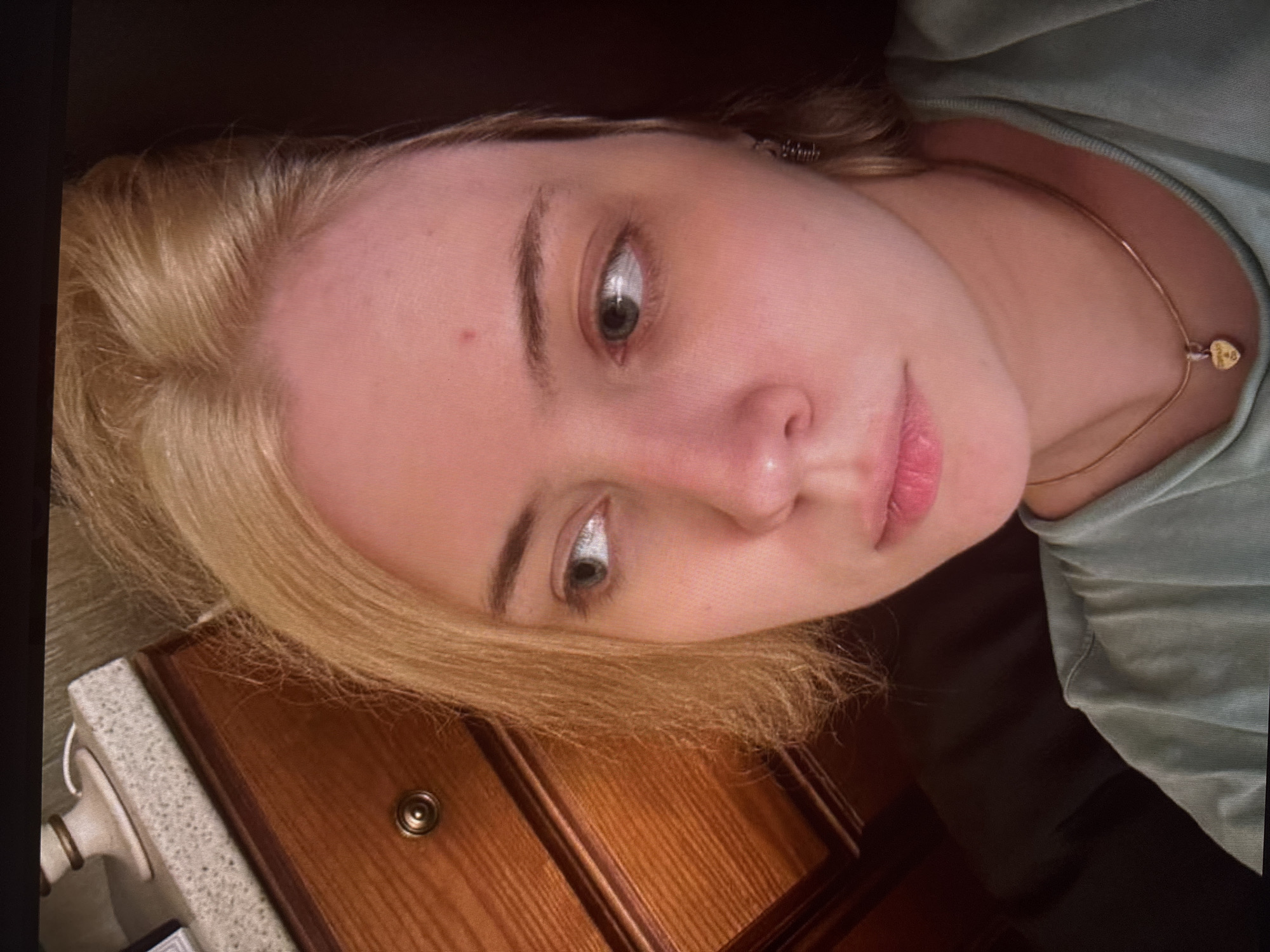 A young Caucasian female with blonde hair is looking to her left, with her head angled downwards. She is wearing a light green t-shirt and a gold necklace with a heart-shaped pendant. Her skin is pale with some redness on her cheeks and forehead. She has green eyes and light brown eyebrows. Her lips are slightly parted. In the background, there is a wooden dresser with a drawer knob and a portion of a countertop with a lamp. The scene is indoors, with a dark background.