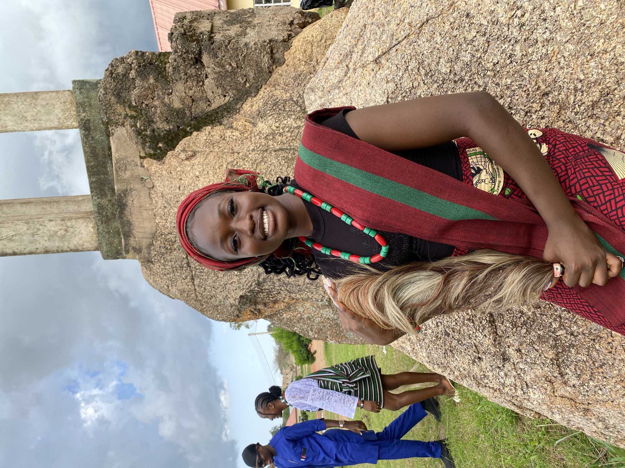 The image shows three people in an outdoor setting with large rocks and grassy ground. A young woman is prominently featured in the foreground, leaning against a rock. She has dark skin, dark curly hair, and is wearing a red headscarf. She is smiling at the camera and has a beaded necklace of red and green beads. Around her shoulders, she wears a sash or fabric with a red background and green stripes. Her right arm is visible, and she appears to be holding something light-colored, possibly hair extensions, in her hand. A ring is visible on her right index finger.
In the background, two other individuals are partially visible. One person, wearing a white top and a striped skirt, is looking down. Another person, wearing a blue outfit and sunglasses, is also visible.
The scene appears to be in a rural or natural location, with large, weathered rocks dominating the landscape. The sky is cloudy, suggesting it might be overcast or that rain could be approaching. A building with a pink roof is visible in the upper part of the image. The presence of people in traditional or festive attire and the natural setting suggest a gathering or cultural event. No visible text is present in the image. The overall mood seems cheerful, primarily conveyed by the smiling expression of the woman in the foreground.