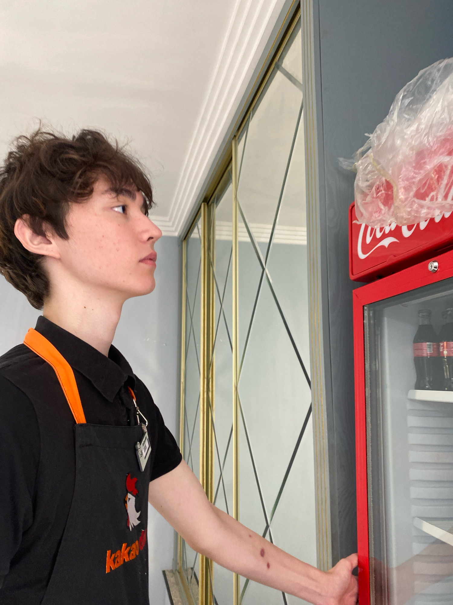 A young male, possibly of East Asian descent, with brown, wavy hair, is seen in profile, looking to the left. He is wearing a black collared shirt with orange straps and a black apron with an embroidered chicken logo and orange lettering that reads "kakariki". He has a small, square, grey tag attached to the apron. His right arm is extended towards a red Coca-Cola branded refrigerator, which is partially open. The refrigerator contains several bottles of Coca-Cola. The background features a mirrored wardrobe with diamond-shaped panels and a grey wall. The lighting suggests an indoor environment, possibly a kitchen or a retail space. The image does not provide any specific clues about its location in the city of Astana, Kazakhstan.