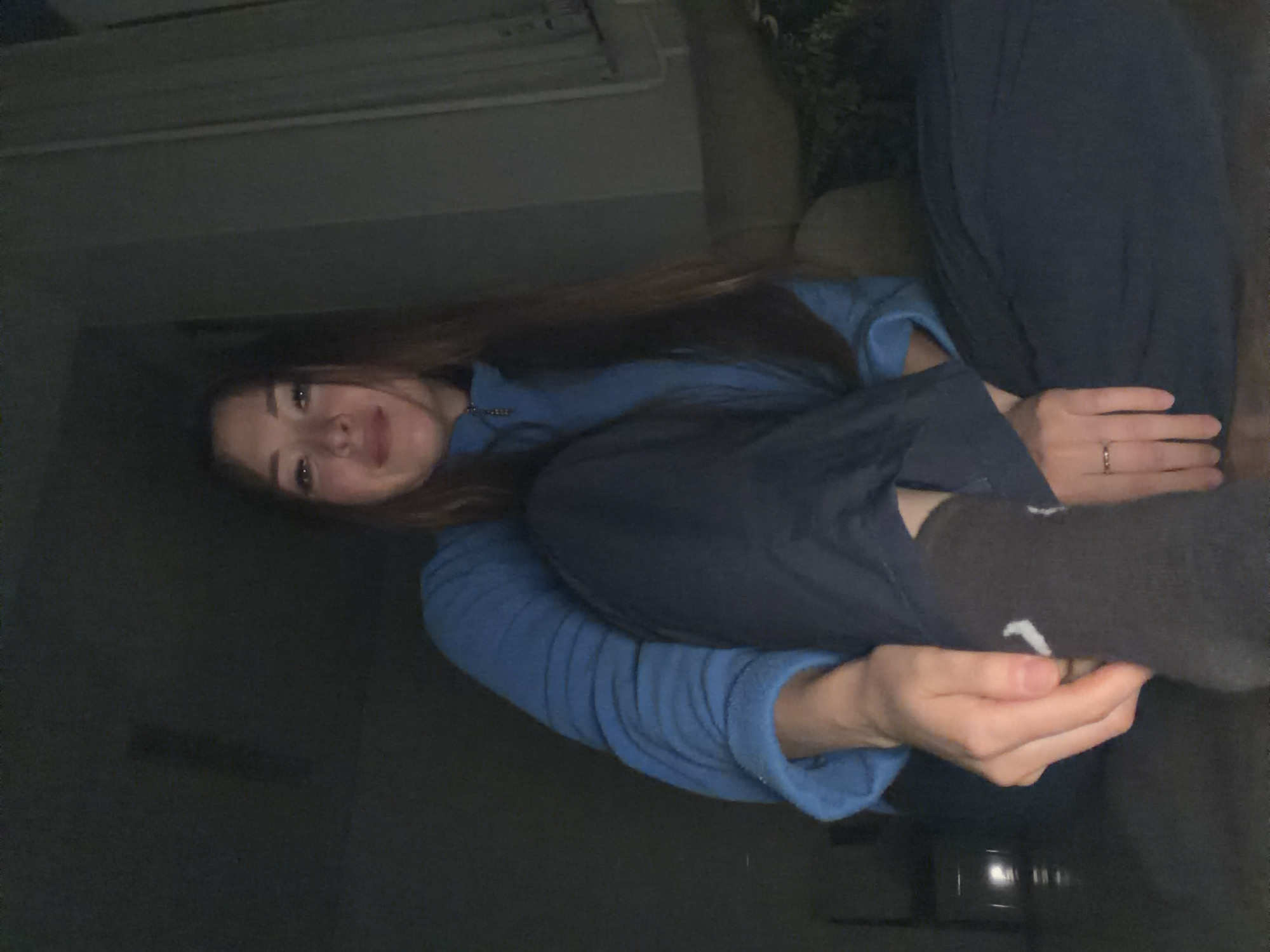 A young woman is sitting on the floor in a dimly lit room. She is wearing a blue fleece jacket and dark pants. Her legs are crossed, and she is resting her hands on her right leg. The room appears to be a living room, with a sofa visible in the background. The overall scene is casual and relaxed. There is no information in the image to suggest this location is in the city of Япрыково, Russia.