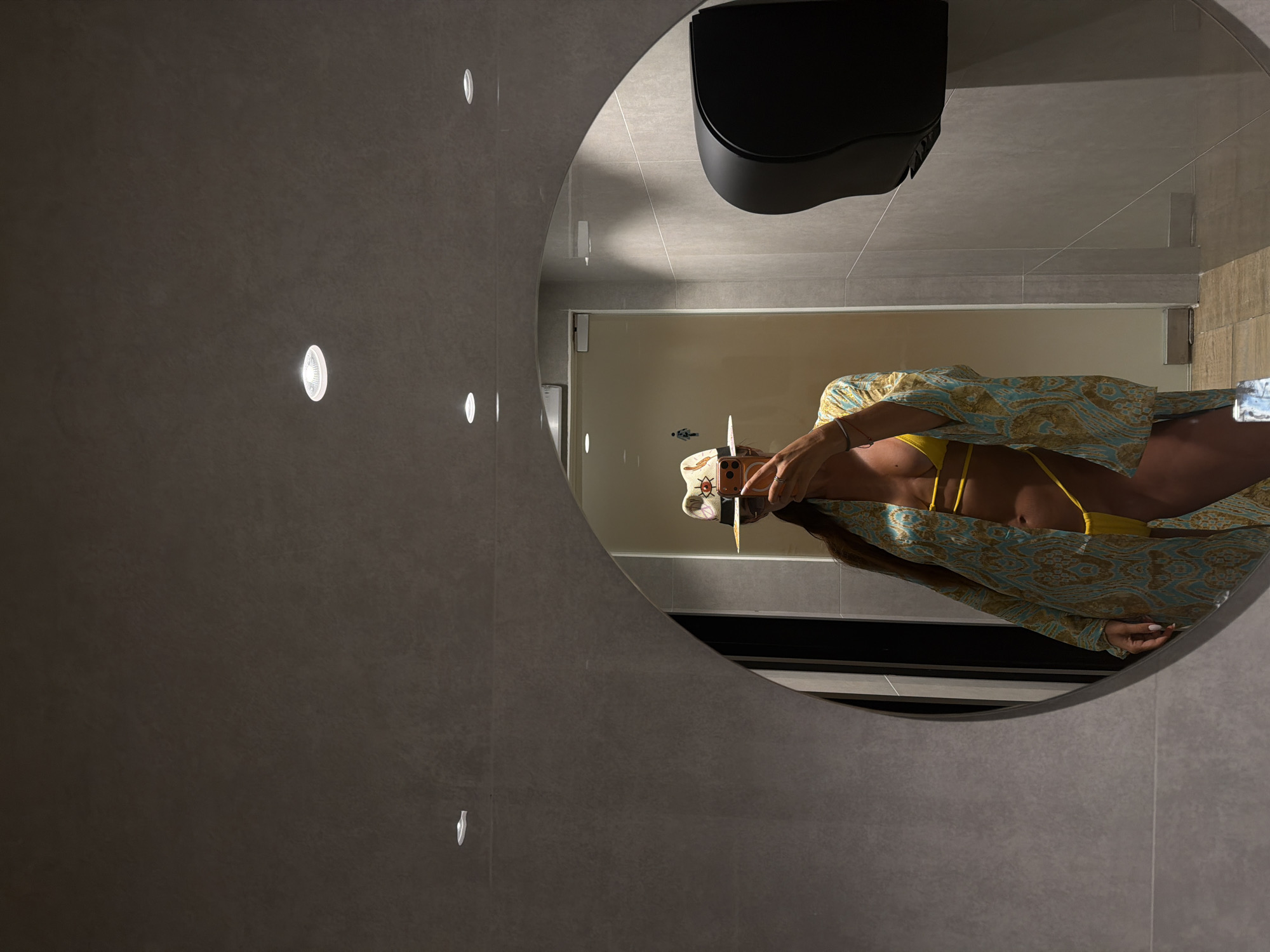 A person wearing a yellow bikini and a patterned kimono takes a mirror selfie in a modern restroom. The person is also wearing a straw hat with a colorful band. The restroom features gray tiled walls, a large oval mirror, and a black paper towel dispenser. The lighting is dim, with recessed ceiling lights providing illumination. The floor is tiled with a lighter brown material. The overall impression is one of a private or hotel restroom.