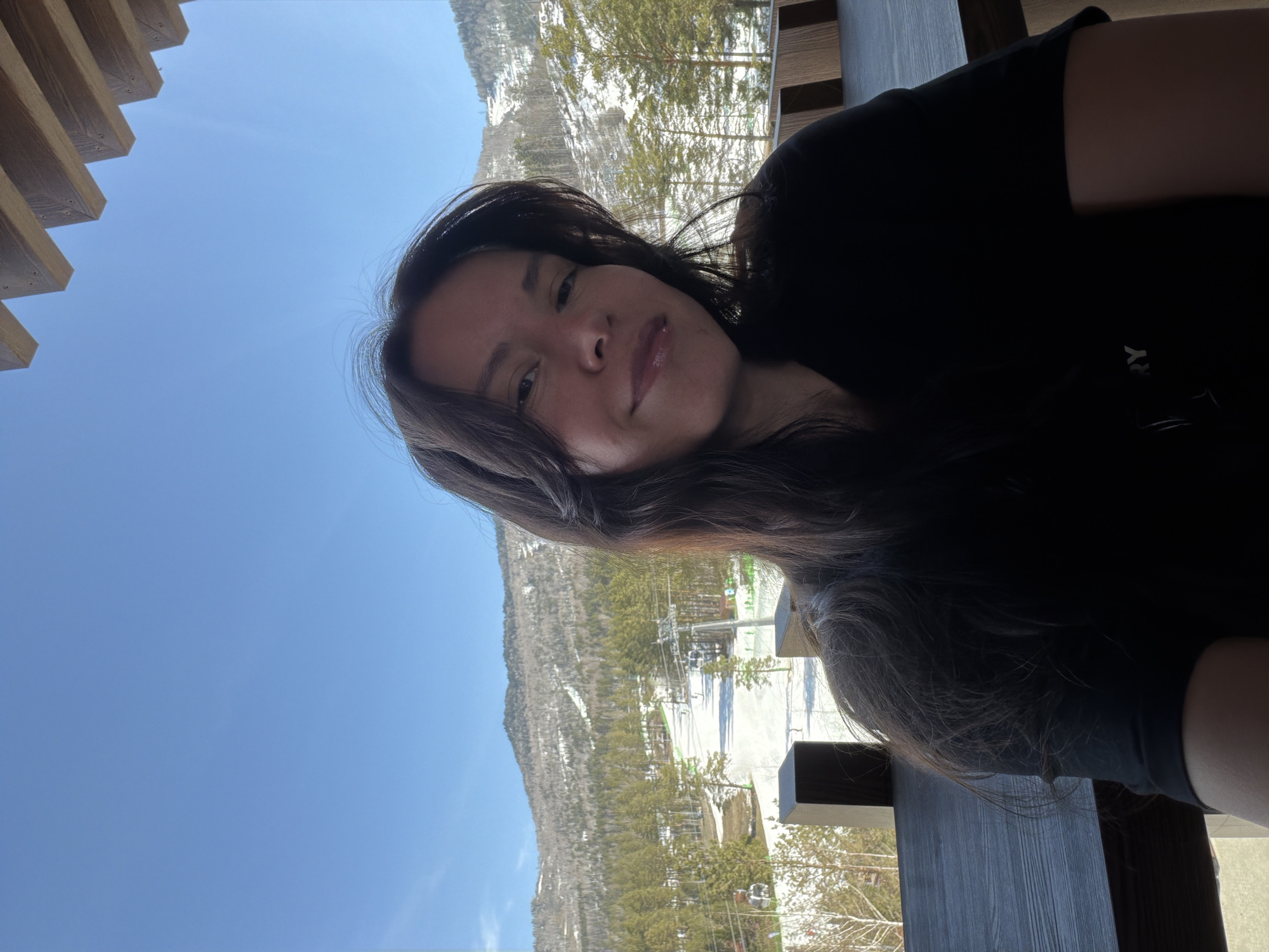 A selfie-style photograph shows a person with long dark hair, presumably female, facing the camera with a slight smile. The person is wearing a black shirt. The background reveals a mountain landscape, likely a ski resort, with snow-covered slopes and trees. In the distance, ski lifts are visible. The sky is clear and blue. A wooden structure, possibly a balcony or deck, is in the foreground. The location appears to be in a mountainous region, possibly near Озёрное, Russia.