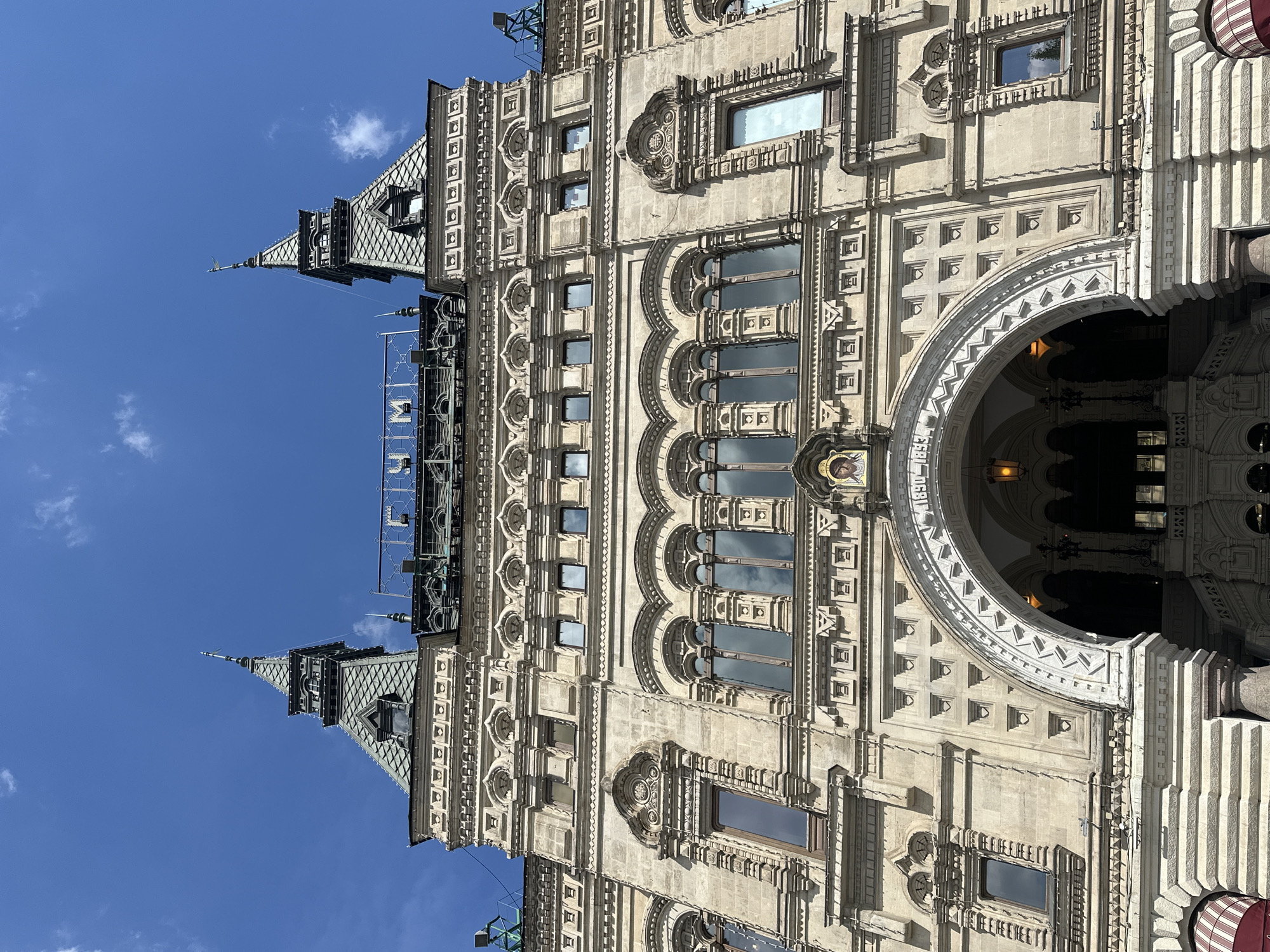 This is a low-angle shot of the facade of a grand, ornate building, likely a historical landmark, under a clear blue sky with scattered white clouds. The architecture is detailed and elaborate, featuring multiple stories with numerous windows, decorative carvings, and arched entrances. The building's material appears to be stone, with a light beige or cream color.
Notable architectural elements include pointed spires and turrets on the upper sections, characteristic of older European buildings. The facade is rich with ornamental details such as intricate molding, recessed panels with geometric patterns, and round decorative elements above some windows. A large, semicircular archway is visible, leading into a darker interior space. Above this arch, text is partially visible, reading "1890".
The sunlight suggests it is daytime, likely late morning or early afternoon, casting subtle shadows that highlight the building's three-dimensional features. The overall scene conveys a sense of historical grandeur and architectural splendor. The image is oriented vertically, with the sky occupying the left side and the building the right. The clear sky and sunlight indicate fair weather. No people are visible in the frame, making the building the sole subject. The context provided indicates the location is Moscow, Russia.