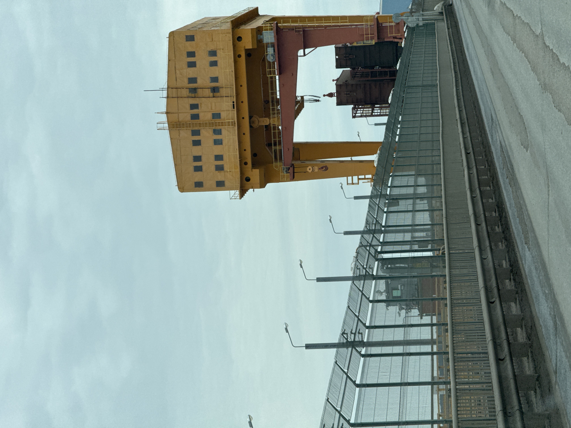 A yellow gantry crane dominates the scene, its boxy cabin featuring multiple window openings. The crane's boom extends toward the right of the frame, with some rust-colored elements. A metallic fence with light fixtures runs horizontally in the foreground, partially obscuring the lower portion of the crane. The background consists of a gray sky. The image is rotated counter-clockwise. Without any information about landmarks in Chaikovsky, Russia, to use as identifiers, the location cannot be pinpointed.