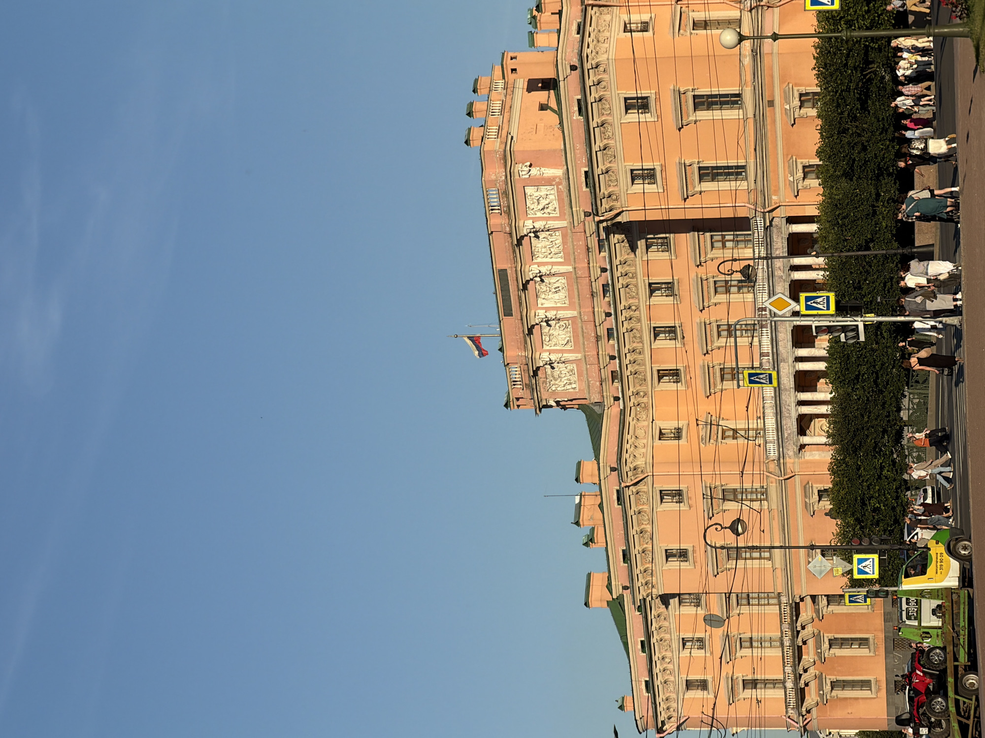 The image shows a street scene in Saint Petersburg, Russia, in front of a large, ornate, peach-colored building. The building has multiple stories, a green roof, and decorative architectural elements, including sculpted friezes. A Russian flag is visible flying from the top of the building. The sky is clear and blue.
In the foreground, a street with a crosswalk is visible. Several people are crossing the street. On the left side of the street, a tow truck is parked, with a red ATV on its trailer. There are traffic lights and pedestrian crossing signs at the intersection. A lamppost with a decorative lantern stands on the sidewalk. Dense green hedges line the front of the building.