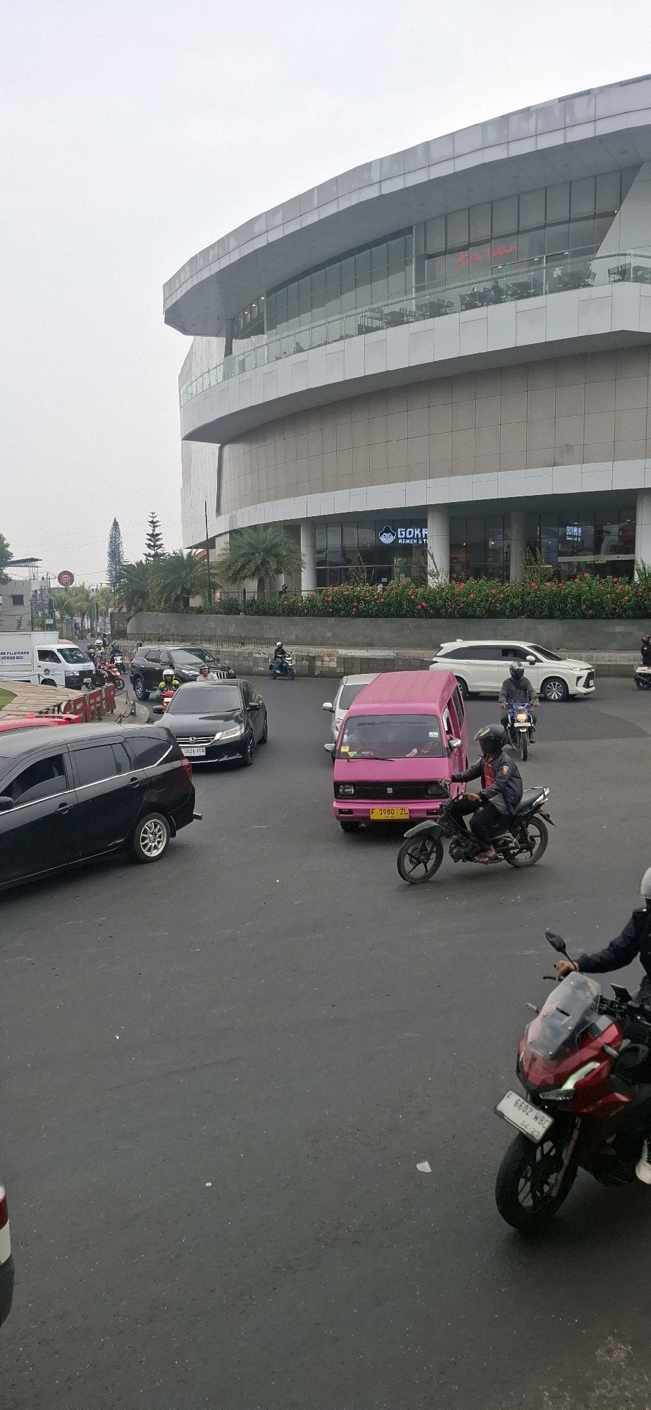 This is a street-level, eye-level, slightly high-angle, full-shot photograph taken on a street in the city of Cianjur, Indonesia. The primary subject is a busy intersection in front of a modern, multi-story building with a curved, gray facade and large glass windows. The building appears to be a commercial establishment, possibly a mall or office building, with visible signs of businesses like "GOKRA REMEN & YOGA" on the ground floor.
Several vehicles are present, including cars and motorcycles, indicating traffic flow. In the foreground, a black sedan and a black SUV are stopped at the intersection. A pink van is positioned to the right of the sedan, with a motorcycle rider in black clothing and a helmet in front of it. Another motorcycle, this one red, is in the lower right corner, with its rider also wearing dark clothing and a helmet, facing away from the camera. Several other motorcycles are visible throughout the scene, some with riders and others parked or moving.
The environment appears to be urban, with paved roads and sidewalks. There are some trees and greenery visible, particularly around the base of the modern building. The sky is overcast and a light gray, suggesting it might be a cloudy or hazy day. The lighting is diffused, with no harsh shadows. The overall impression is of a typical day in a bustling Indonesian city.
