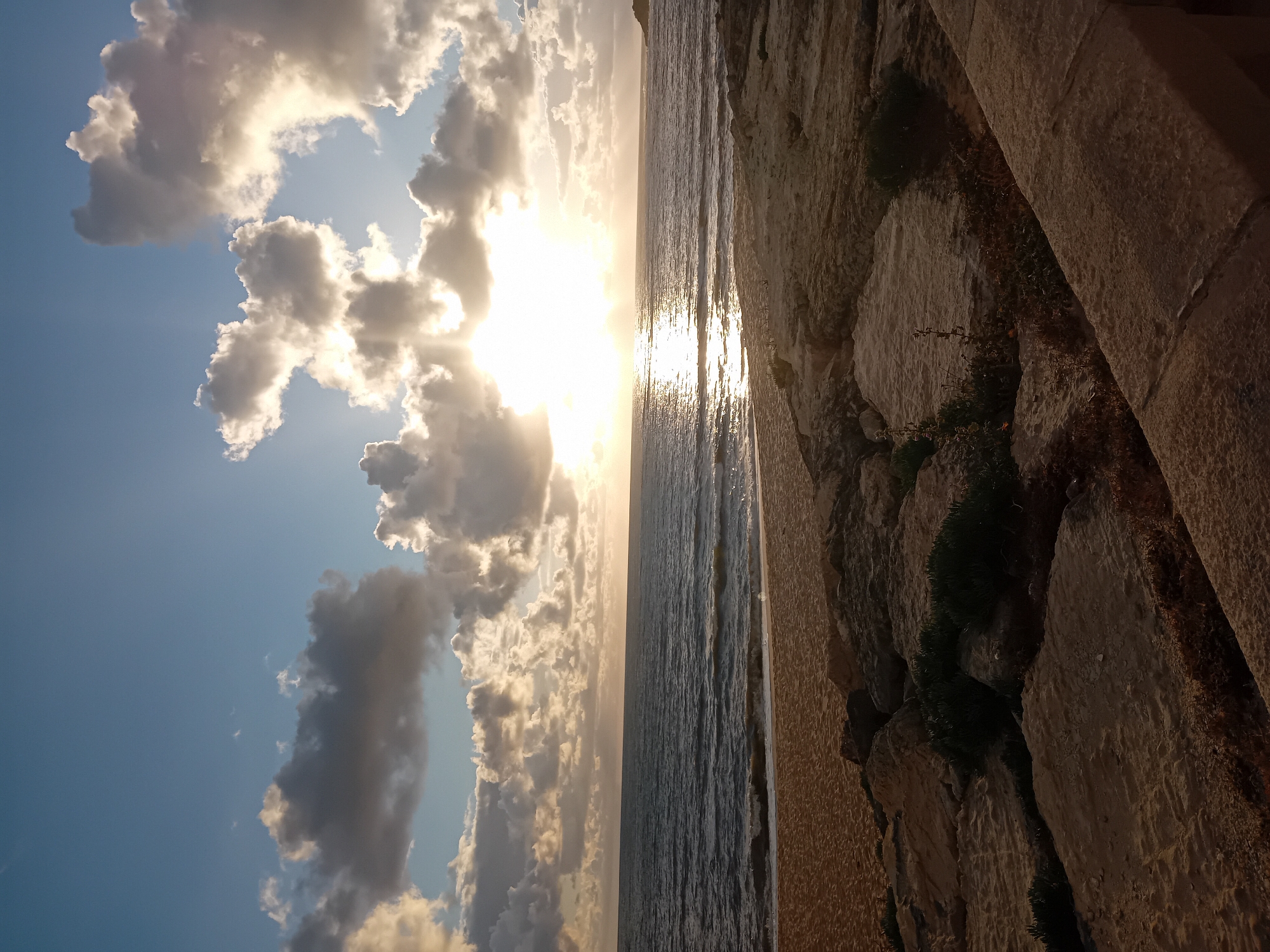 The image depicts a view of the ocean at sunset, possibly from a cliffside. The sky features scattered clouds illuminated by the setting sun. The ocean's surface reflects the sunlight, creating a bright streak. A wall or cliff face is visible on the right side of the image, composed of textured stone and vegetation. The location is Unknown, Unknown.