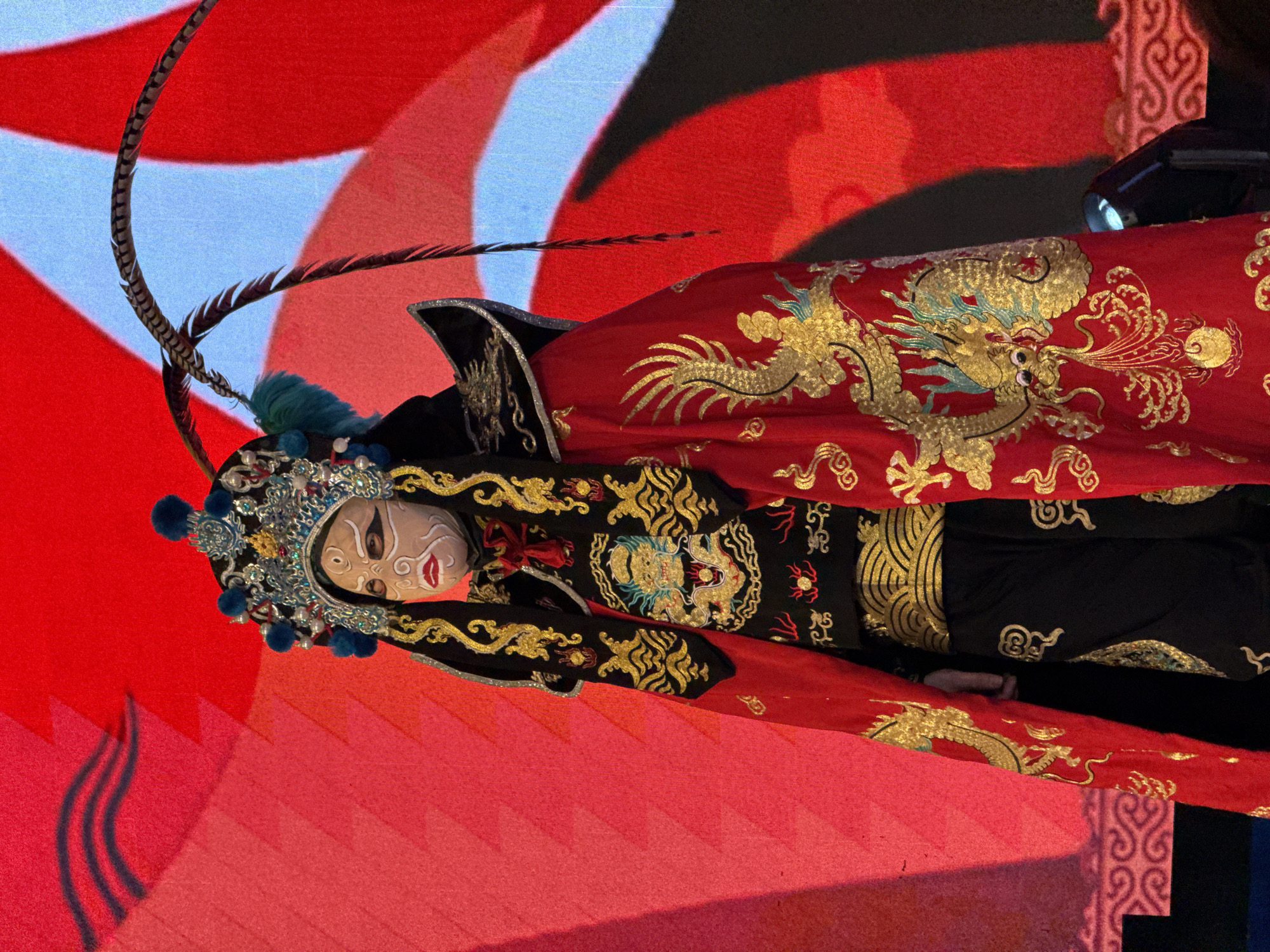 The image displays a person or a highly realistic mannequin adorned in an elaborate traditional Chinese opera costume, likely Peking Opera, set against a brightly lit, theatrical backdrop. The figure is positioned slightly off-center to the left, shown from approximately the waist up.
The central subject wears an ornate, multi-layered headpiece, rich with silver-toned metalwork, blue and red jeweled accents, and prominent blue pom-poms. Long, slender pheasant feathers, characteristic of opera attire, curve elegantly upwards and to the right from the headwear. The face is painted in a stylized, mask-like fashion: a pale white base with dramatic black outlines for the eyes and brows, striking red lips, and subtle red detailing on the cheeks and forehead.
The costume itself is highly decorative, featuring a combination of black and vibrant red fabrics. A voluminous red outer garment is lavishly embroidered with intricate golden dragon motifs, depicted with scales, claws, and swirling patterns, suggesting movement and power. Beneath this, a black inner garment is visible, also adorned with detailed gold embroidery, including stylized patterns and smaller mythical creatures. A gloved hand, matching the costume's aesthetic, can be seen at the lower edge of the frame.
The background is a dynamic composition of large, abstract shapes in bold red and lighter blue hues, resembling stylized clouds or flames, possibly projected onto a screen or part of a stage design. The lighting is bright and artificial, typical of an indoor performance or exhibition setting. There is no visible text in the image. The overall scene evokes cultural richness and dramatic presentation, characteristic of a performance or display in Yuzhong District, China.