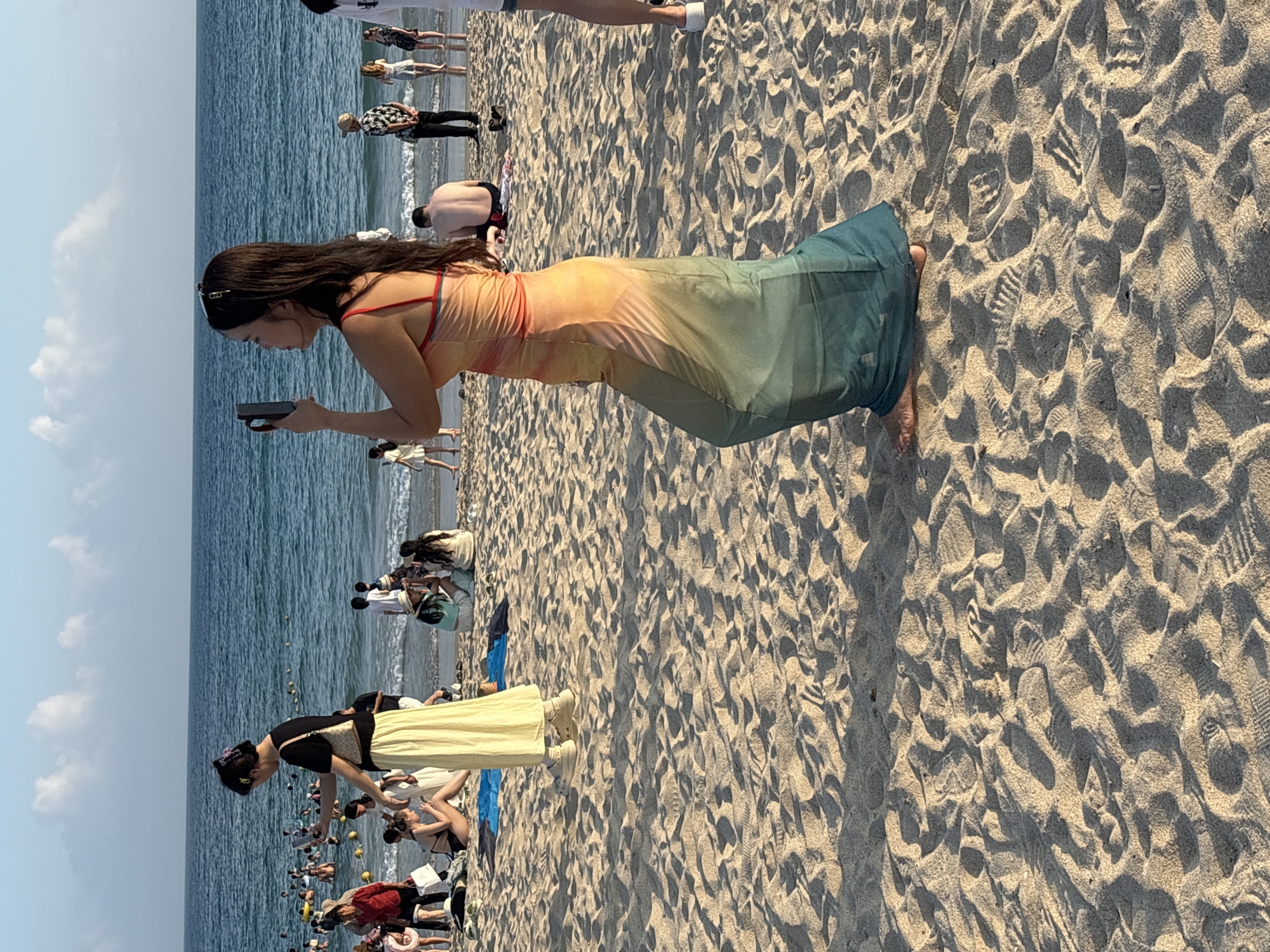 This is a photograph taken on a crowded beach in Jiyang District, China. The scene is dominated by sand, with the ocean visible in the background. The sky is a pale blue with scattered white clouds. The water is calm and appears to have some buoys or markers in it, indicating a designated swimming area.
In the foreground, a woman is captured in mid-stride, holding a smartphone up as if to take a photo or video. She is wearing a long, flowing dress that transitions in color from orange at the top to a teal green at the hem. She has long, dark hair and appears to be looking off to her right.
Behind her, other beachgoers are scattered across the sand and in the water. To the left of the woman, another woman in a black top and a long, pale yellow skirt is also holding a smartphone, seemingly engaged with it. Further back and to the left, a large group of people are visible in the shallow water, enjoying the sea.
The sand itself is textured, showing footprints and undulations from the tide and the movement of people. The lighting suggests it is either late afternoon or early morning, casting long shadows across the beach. The overall impression is of a lively and populated beach scene.