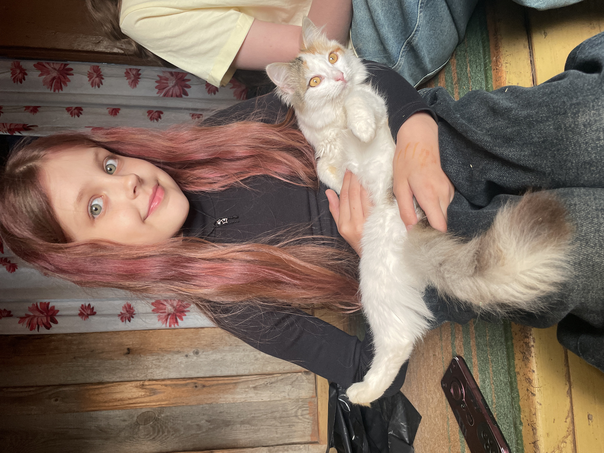 A girl with long, light auburn and pink hair, a fair complexion, and light-colored eyes is holding a white and light brown cat. The cat is in a reclined position on its back. The girl is wearing a black long-sleeved shirt. Another person is visible above them wearing a yellow shirt and blue denim. They appear to be seated. The background consists of a wooden plank wall with a floral print curtain above. A portion of a cell phone and a black plastic bag are visible on the lower right. The location is in Балдейка, Russia.