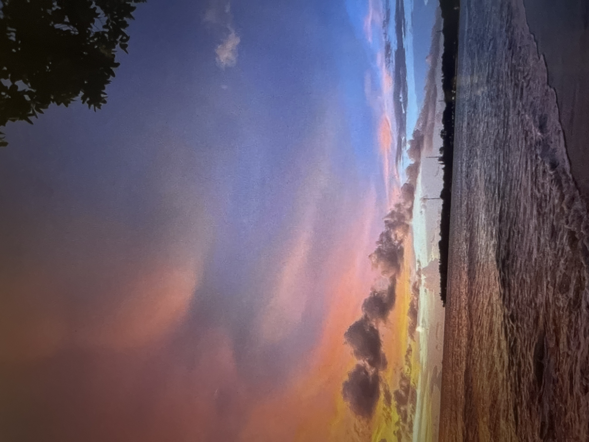 The image depicts a coastal scene at dusk. The sky is filled with vibrant colors, transitioning from orange and yellow near the horizon to shades of pink, purple, and blue higher up. Several clouds are visible, some dark and textured, others wispy and illuminated by the setting sun. The ocean is calm, with gentle waves lapping at a sandy shore in the foreground. A dark silhouette of trees and structures can be seen along the distant coastline, hinting at a populated area. The overall atmosphere is serene and colorful, characteristic of a sunset over the water.