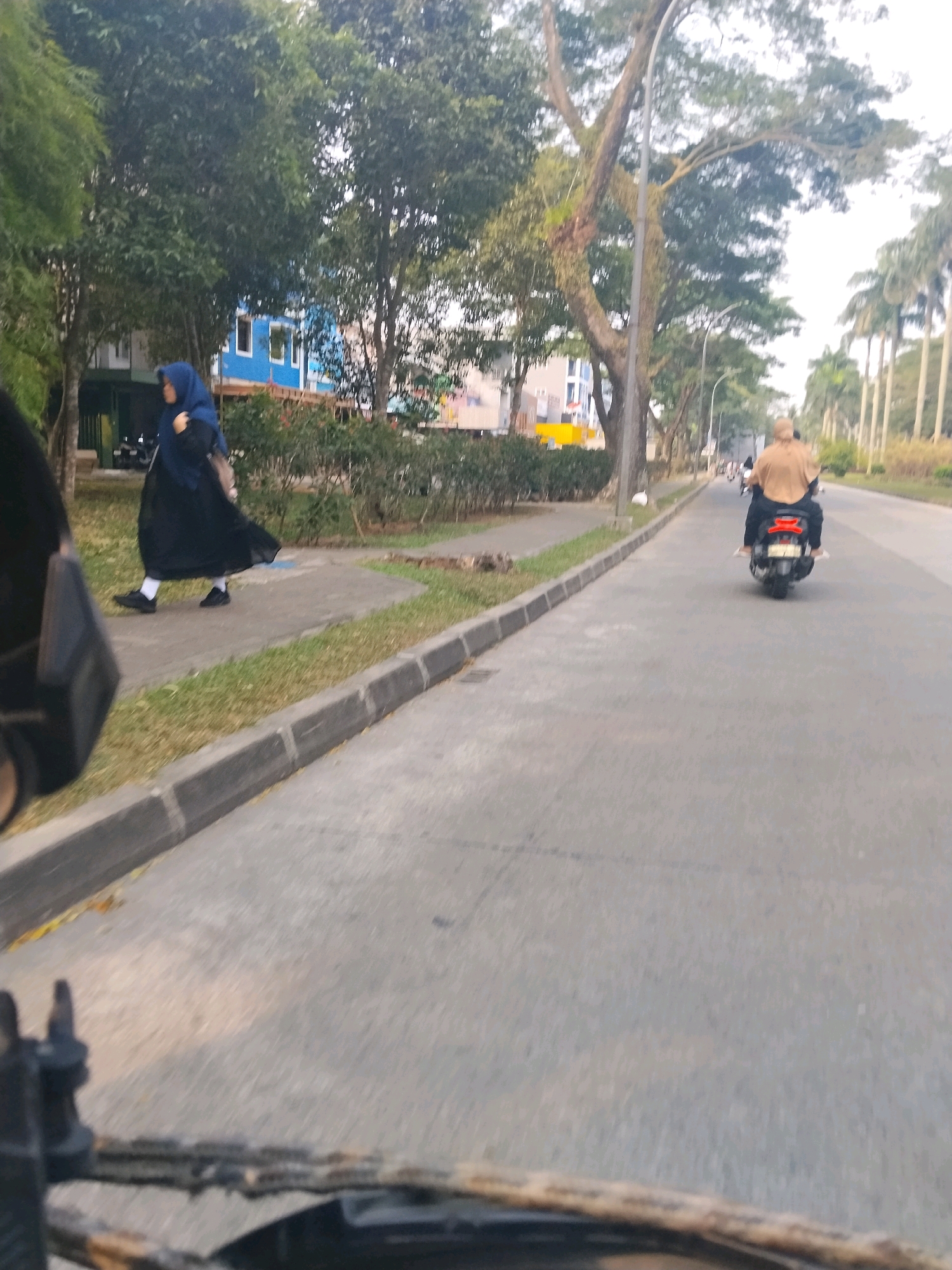 In Cikupa, Indonesia, a woman wearing a blue hijab and a long black dress walks on a sidewalk next to a road. A person wearing a tan hijab and a tan top rides a motorcycle on the road, facing away from the camera. Palm trees line the side of the road. The scene appears to be a street in a suburban or residential area of Cikupa.