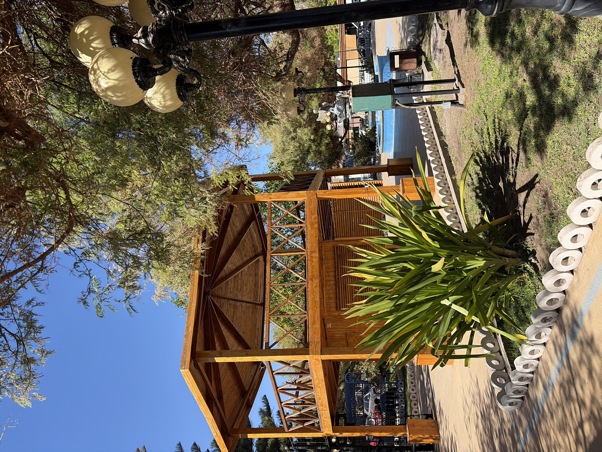 This image captures a sunny outdoor scene in the city of Pica, Chile. In the foreground, a large, lush green plant with long, sword-like leaves is prominently displayed. It is bordered by a decorative line of circular concrete stones. Behind the plant, a two-story wooden gazebo with a lattice-like railing stands out. Its wooden structure is visible, with a wooden roof overhead. The gazebo is situated on a paved area.
Further in the background, a blue fountain with a decorative statue is visible, suggesting a public park or plaza setting. A lamppost with multiple light fixtures is present on the right side of the image, partially obscuring some of the scene. Several trees with dense foliage provide shade and contribute to the natural ambiance of the location. The sky above is a clear, vibrant blue, indicating fair weather. In the distance, vehicles are parked, and glimpses of buildings and fences can be observed. The overall impression is one of a well-maintained public space, likely a park or a recreational area, in Pica, Chile.