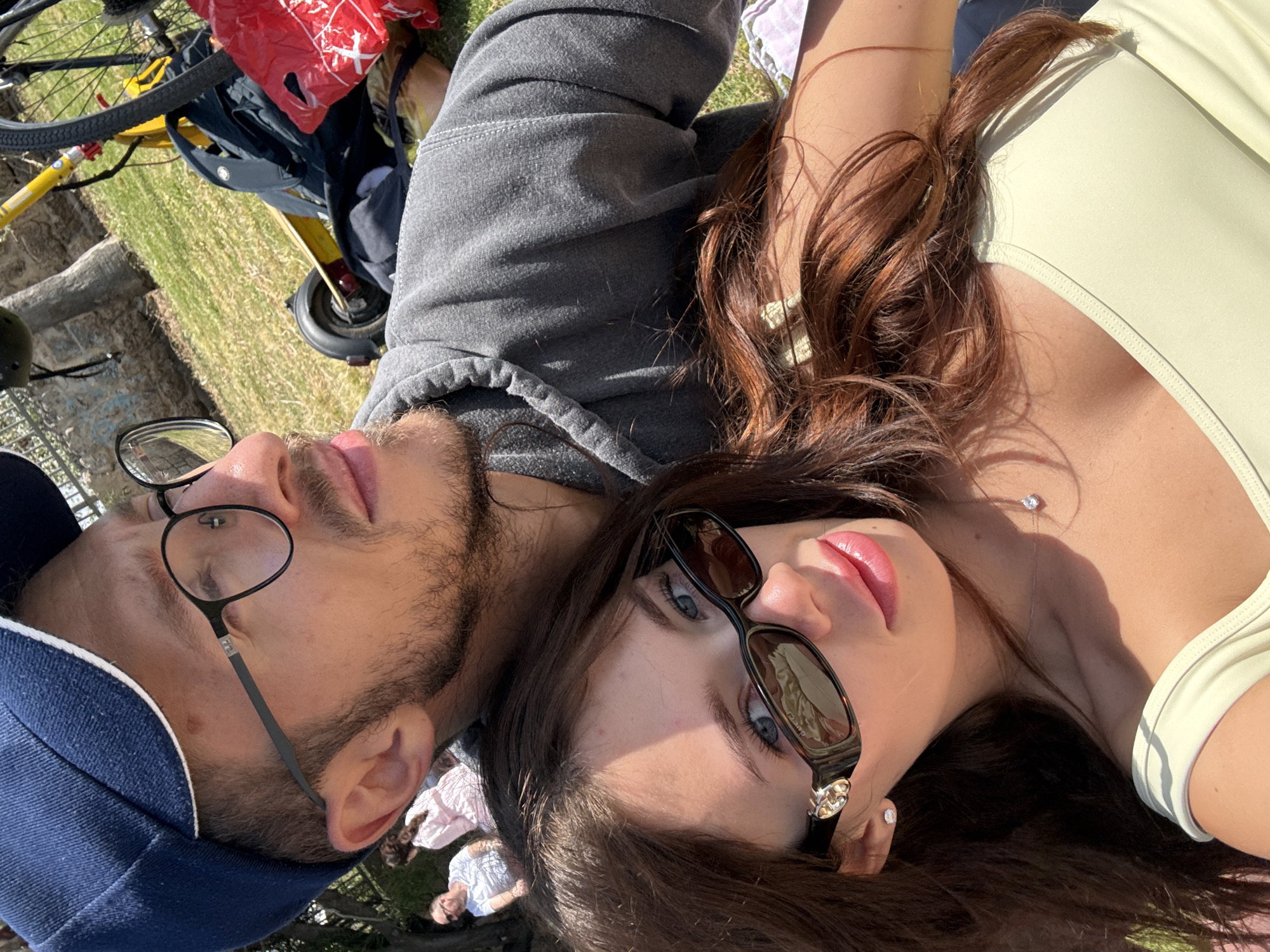 Two people are lying on grass, presumably in a park. The person on the left is wearing a blue baseball cap and black-rimmed glasses, with a gray hooded sweatshirt. The person on the right is wearing sunglasses and a light green top. A bicycle and bags are visible in the background. Other people are in the far background. The location is likely in a public park in the city of Tel Aviv, Israel.