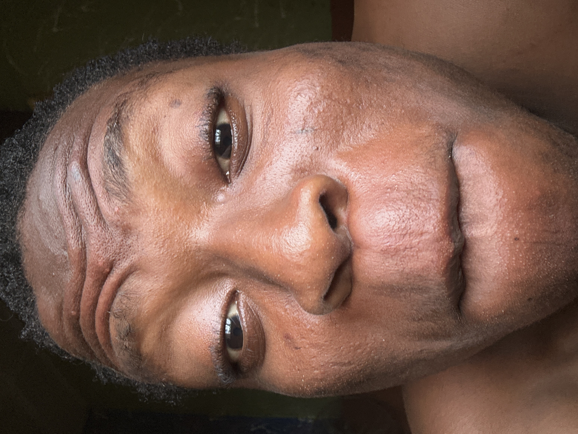 A close-up, slightly angled, portrait of a brown-skinned person. The person has dark eyes, short dark hair, and visible skin pores. The background is dark and indistinct. The location and time are not determinable from the image.