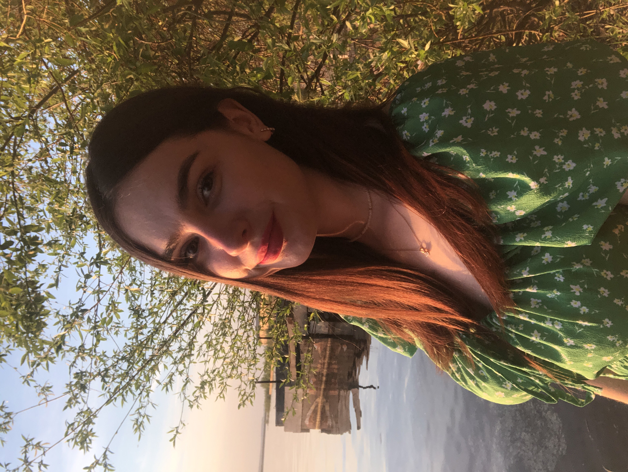 A young woman is the focus of this outdoor photograph. Her face is well-lit with the sun, she has dark hair with some auburn highlights, fair skin, and appears to be wearing lipstick. A gold necklace is visible around her neck. She is wearing a green dress with small white floral patterns. The setting seems to be near water, possibly a lake or river, with reflections of a small wooden structure visible in the water. Trees with green leaves are in the background. This photograph was taken in the city of Chaikovsky, Russia.