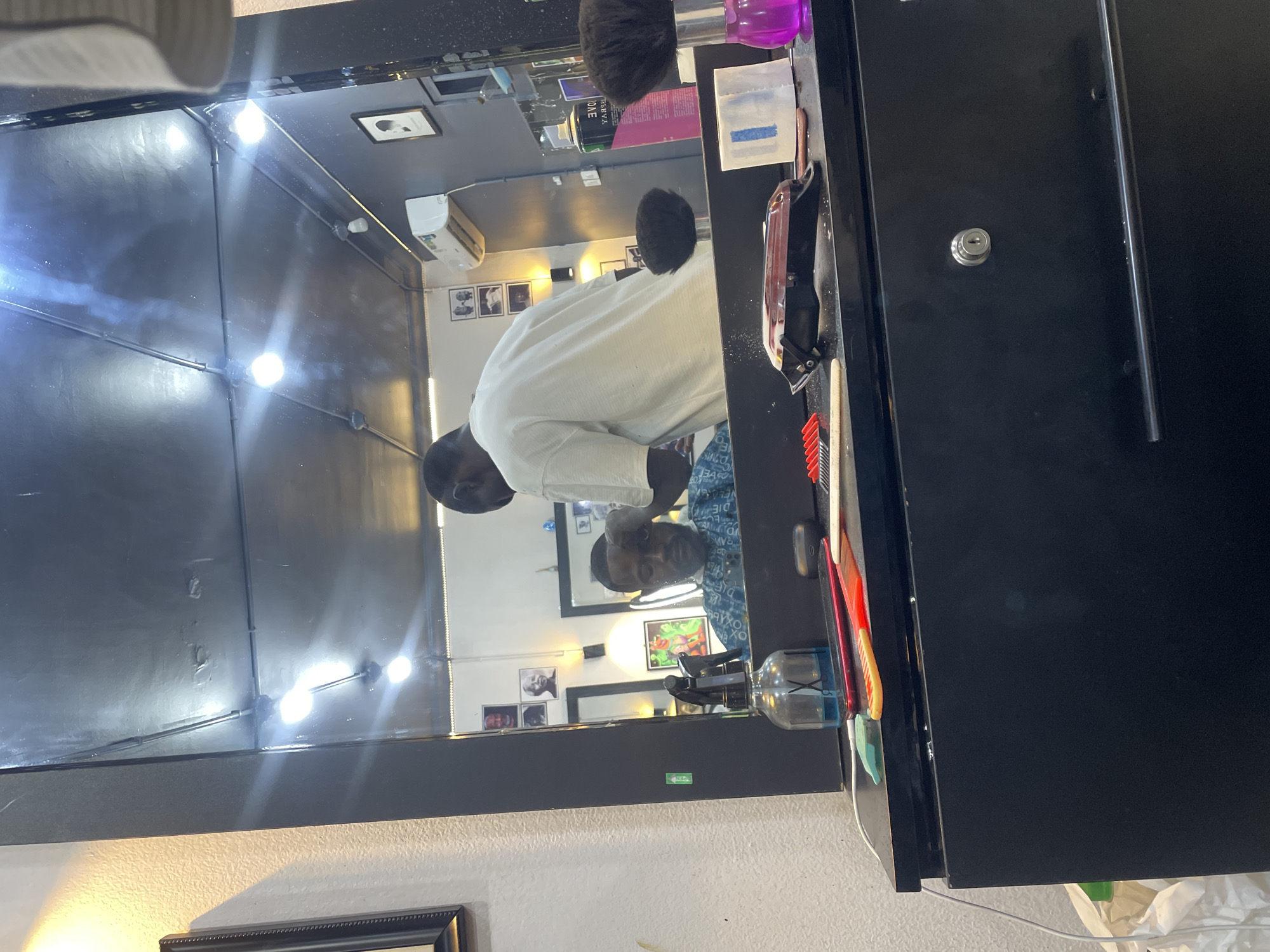 The image captures an indoor scene, likely a barbershop or salon, dominated by a large mirror reflecting the environment. The primary subjects are two individuals visible in the mirror's reflection. One person, presumably the barber, wears a white t-shirt and is seen from the back, leaning over as if performing a service. The second person, the client, wears a patterned blue and white shirt, and their face is visible, looking forward. This suggests a haircut or styling session is underway.
The countertop beneath the mirror holds various barber tools and products. These include hair clippers with a distinctive red and chrome finish, multiple combs (red, yellow, green), a black brush, and several bottles. Visible bottles include one with a purple liquid, another clear bottle with a "X" logo, and a pink spray bottle with the word "TONE" (reversed in reflection). A white square container and charging cables are also present. To the right, a black cabinet-like structure features a visible keyhole.
In the background of the reflection, a dark wall or ceiling is equipped with multiple bright, circular light fixtures that create prominent reflections and lens flares. An air conditioning unit and several framed pictures are also seen on the walls. Below the mirror, a textured white wall is visible, with a framed picture towards the bottom left. A small green sticker or sign is also visible below the mirror's lower edge. The overall lighting is artificial and bright, suggesting an indoor setting without strong natural light cues, making the time of day indeterminate. The image provides a glimpse into a typical barbershop setting in what is contextualized as Abuja, Nigeria.