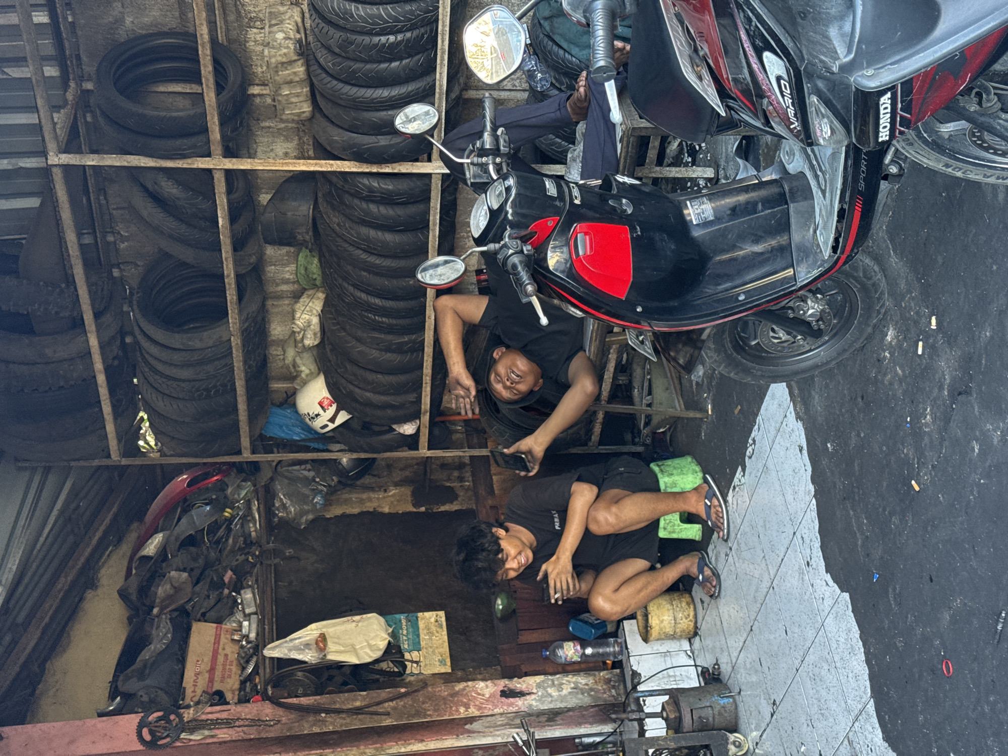 The image depicts the interior of a motorcycle repair shop in Denpasar, Indonesia. The shop is filled with various automotive parts, particularly tires stacked on shelves. Two men are present. One man is seated on a green stool, looking at his phone and holding a cigarette. The other man is lying down on a wooden platform, appearing to be resting. A red and black scooter is parked in the foreground. The environment is cluttered and appears to be a working garage.