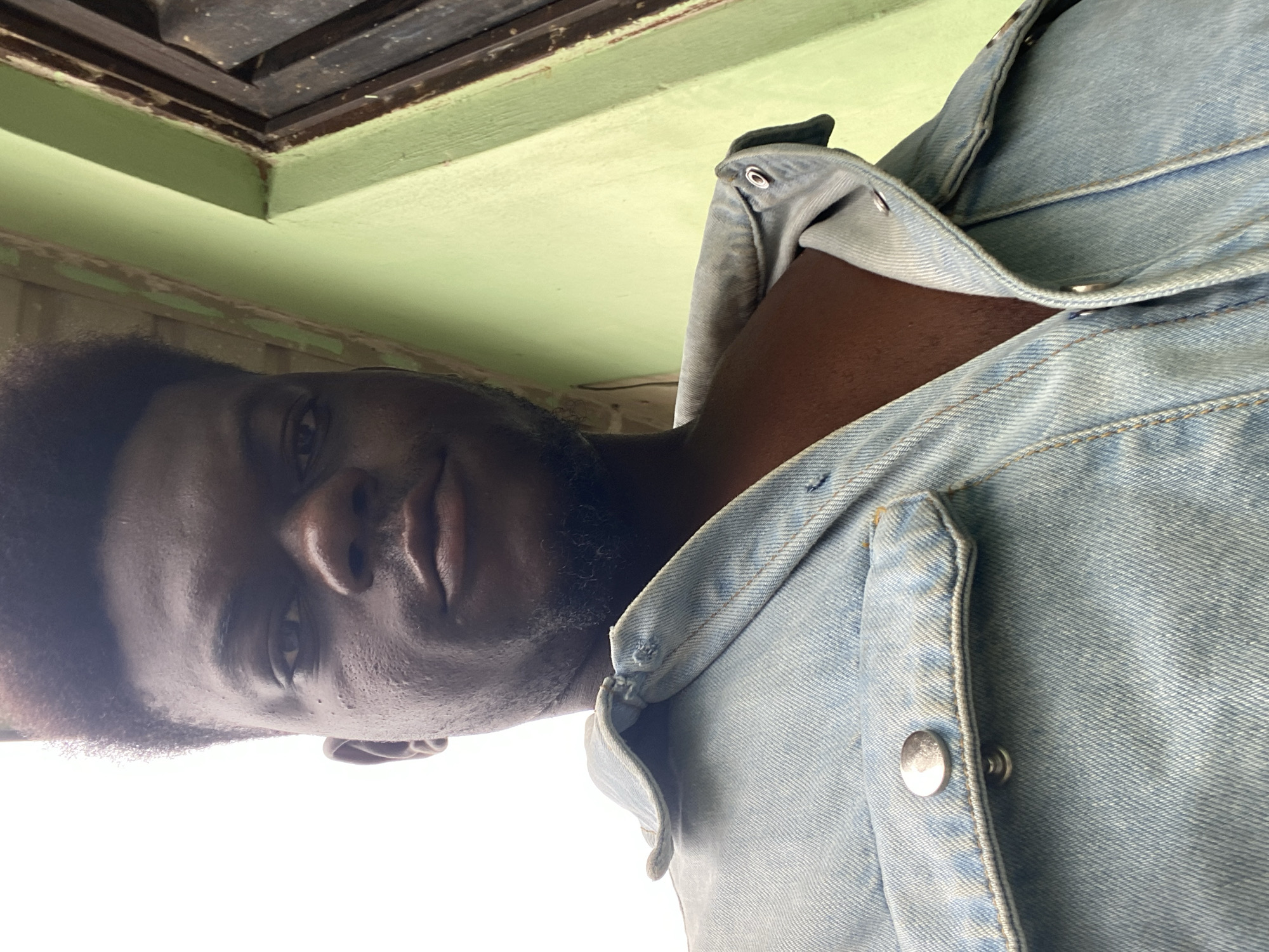 A close-up shot focuses on a person wearing a light-wash denim jacket, with a green wall visible in the background. The person's face is partially visible, showing features such as the eye, nose, and mouth. The denim jacket is detailed with visible buttons and texture. The environment suggests an interior space, possibly a room with a painted wall.
