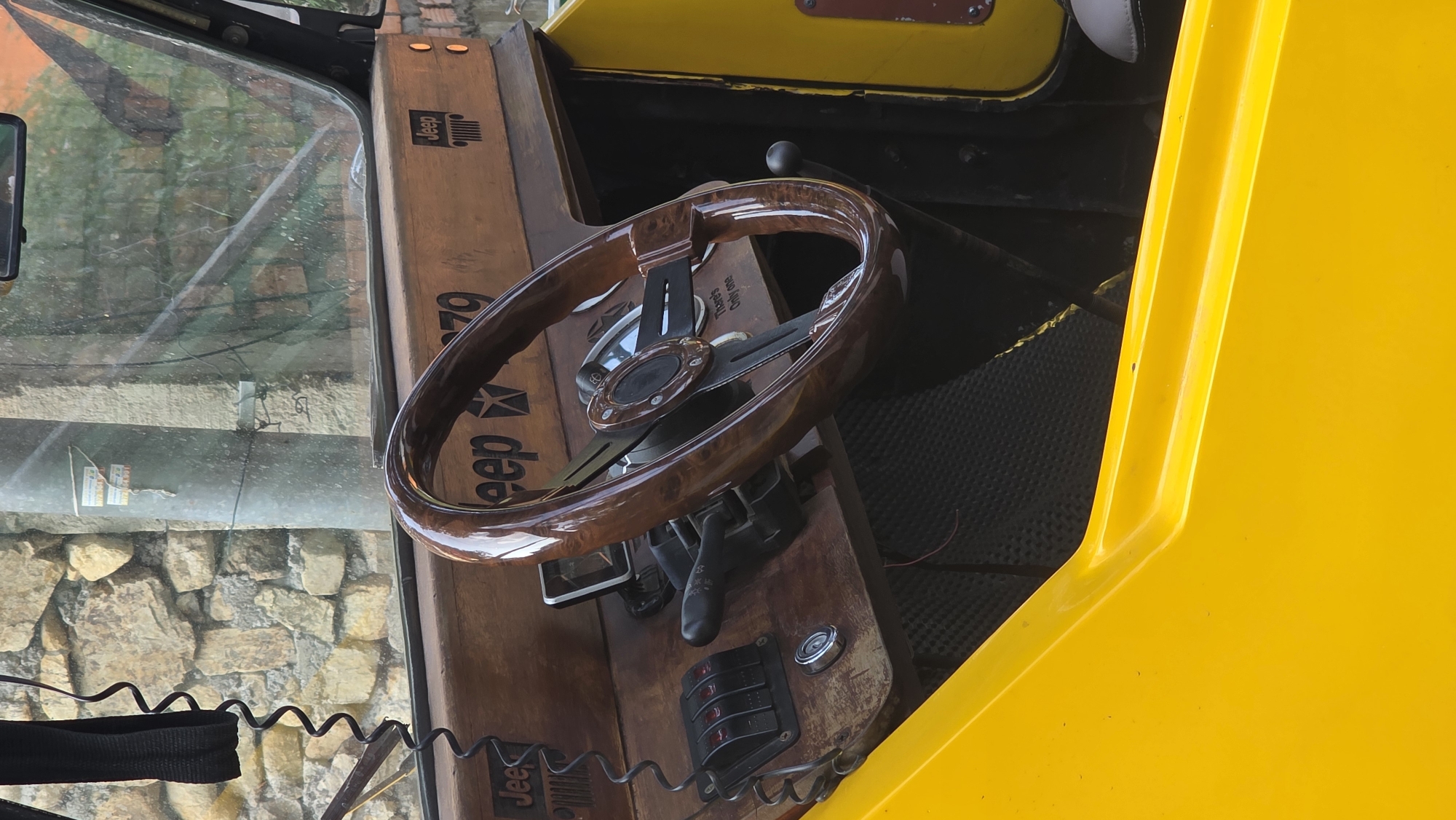 Captured from within a Jeep, likely in the Bac Natural Heritage Area Trang, Vietnam, the image showcases the vehicle's interior. The steering wheel, crafted from polished wood, is a prominent feature. The dashboard, also wooden, displays "Jeep" branding and various gauges and switches. The Jeep's exterior is partly visible, featuring a yellow panel. Through the windshield, a view of a stone or brick structure and vegetation can be seen. A black cable runs across the bottom of the frame. The scene appears to be in daylight.