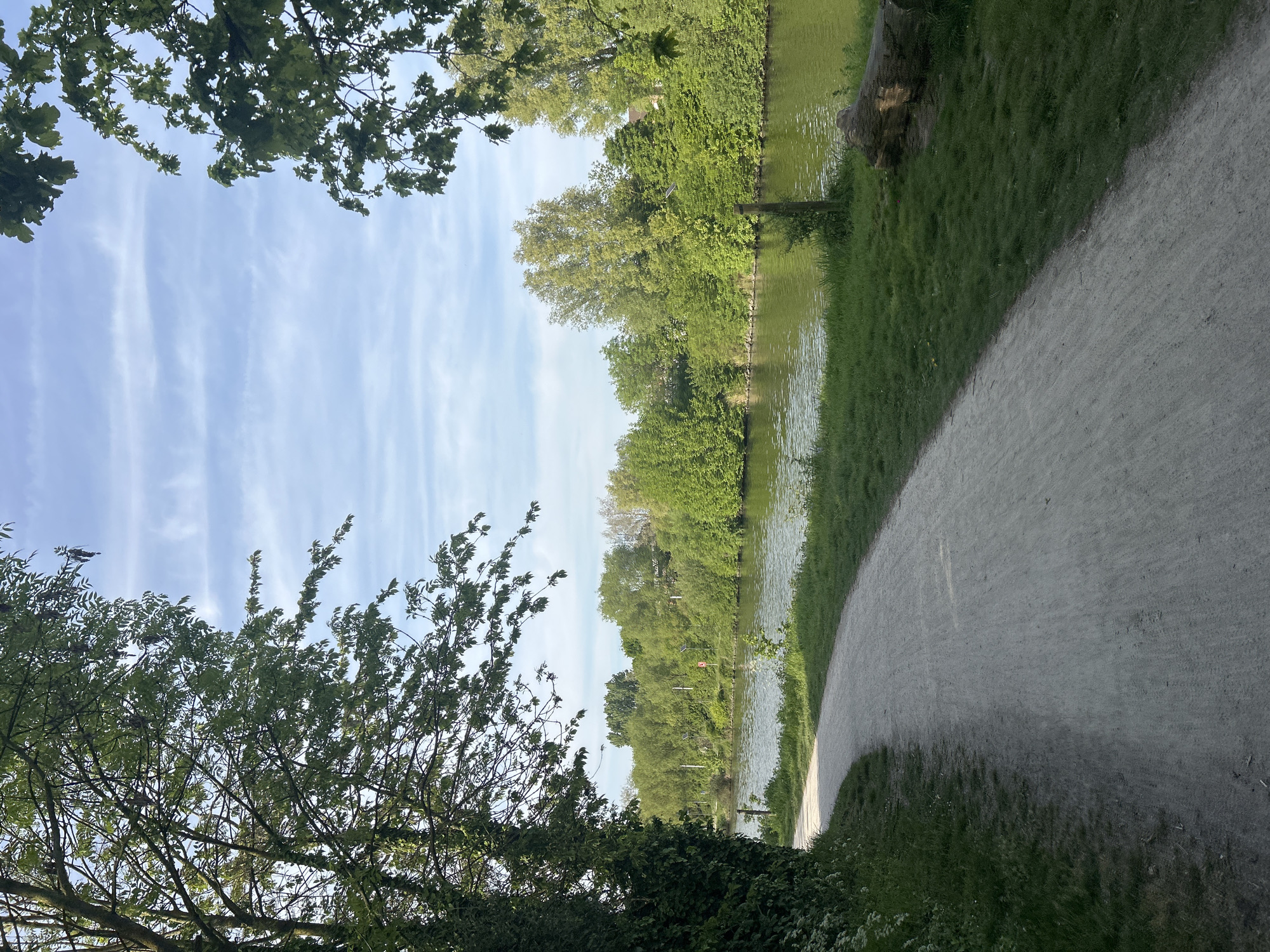 The image depicts a pathway running alongside a body of water, likely a canal or a river, in what appears to be a park or green space. The pathway, composed of light-colored material, curves towards the right. Lush green grass and dense vegetation border the water's edge and the pathway. Trees, with green foliage, frame the top and left side of the image, partially obscuring the sky. The sky is blue with wispy clouds. Based on the location information provided, this scene is located in Marquette-lez-Lille, France. No people are visible in the image.