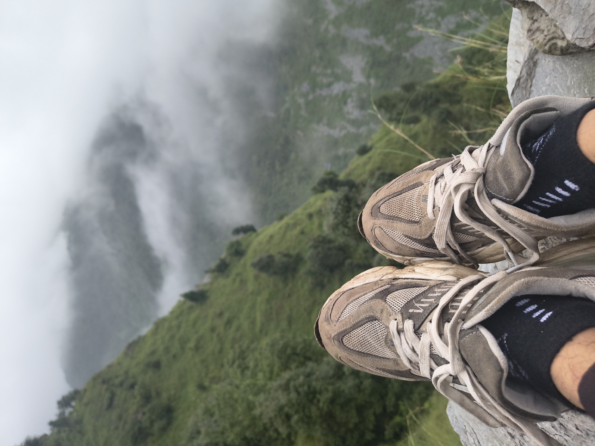 A first-person perspective shot shows a pair of worn, dirt-covered athletic shoes, laced up and resting on a rocky surface. The shoes are a mix of grey and tan with off-white laces. The background is a lush green mountainside, partially obscured by thick fog. The overall scene evokes a sense of being on a hike or trek in a natural, mountainous environment, possibly Bhagsu, India, given the context of the location. The fog suggests a cool, damp atmosphere.