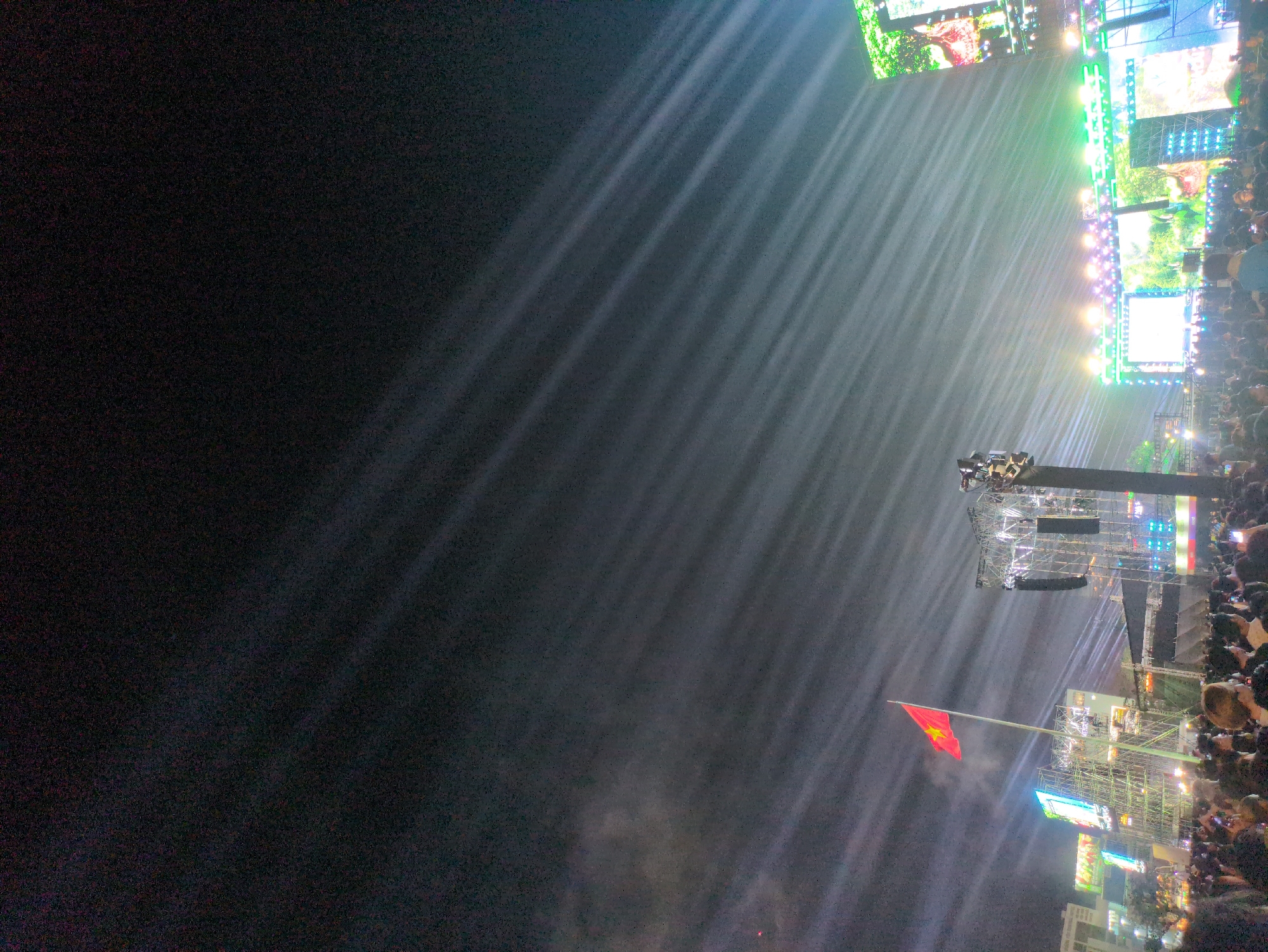 An outdoor nighttime event is depicted in South Quy Nhon Ward, Vietnam, characterized by a large gathering of individuals under a dark sky illuminated by numerous powerful stage lights. The upper two-thirds of the image display a dark sky permeated by parallel beams of white and bluish light extending diagonally from the lower right towards the upper left, indicating atmospheric haze or smoke interacting with spotlights. A faint red light source is visible in the far upper left.
The lower third of the image is occupied by a dense crowd of people, primarily visible from the shoulders up, facing forward towards a large performance area. Many individuals are holding up illuminated smartphones, suggesting active recording or photography.
In the midground, a prominent stage setup is visible. On the right, a large performance stage features multiple brightly lit LED screens displaying vibrant green and yellow imagery, possibly depicting natural scenes or abstract patterns. The stage is framed by horizontal arrays of multicolored lights, including green, blue, and purple. To the left of the main stage, several tall metal truss structures support additional lighting equipment, speakers, and possibly smaller screens. A large Vietnamese flag, red with a yellow star, flies from a pole positioned left of center, above the crowd and in front of a metal support structure, illuminated by ambient light from below. Faint outlines of urban buildings with some illuminated sections are discernible in the far left background.