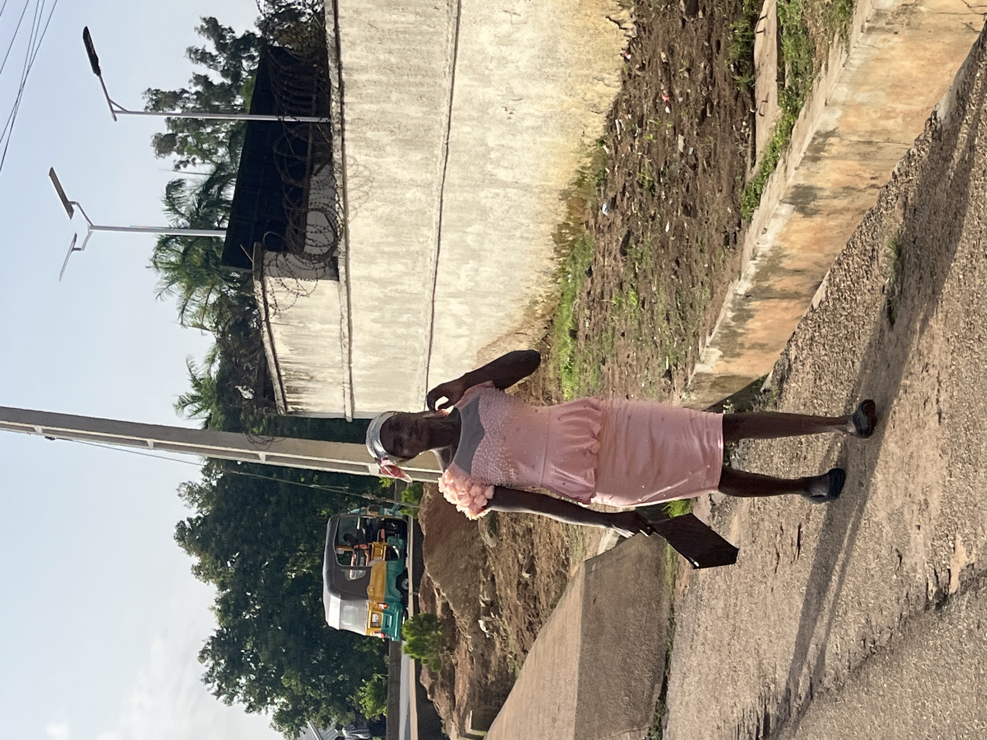 A Black woman walks down a paved street in Bukuru, Nigeria. She is wearing a pink dress and a headscarf. She is carrying a dark-colored bag. In the background, there is a yellow and green tricycle and a wall with barbed wire. The street appears to be in disrepair.