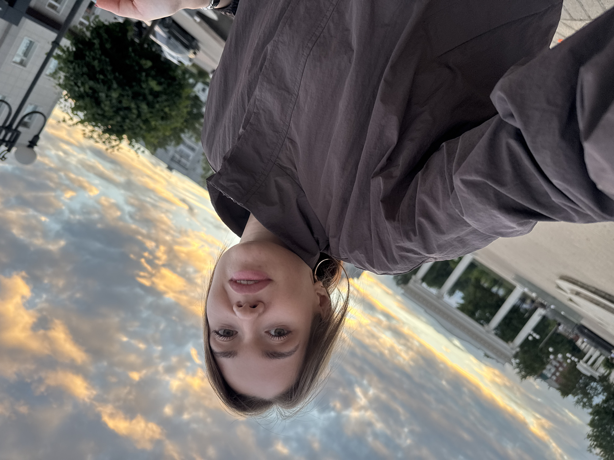 The image is a tilted, close-up selfie of a young woman with shoulder-length brown hair, wearing a dark brown jacket. She has green eyes and is looking towards the camera with a slight smile. She is wearing a large hoop earring in her visible ear. The background appears to be an urban outdoor setting, possibly a park or public square, with architectural elements like columns and trees. The sky above is filled with dramatic clouds, illuminated with warm tones of orange and yellow, suggesting it is either sunrise or sunset. In the distance, a street lamp and buildings are visible. The image was captured in the city of Chaikovsky, Russia.