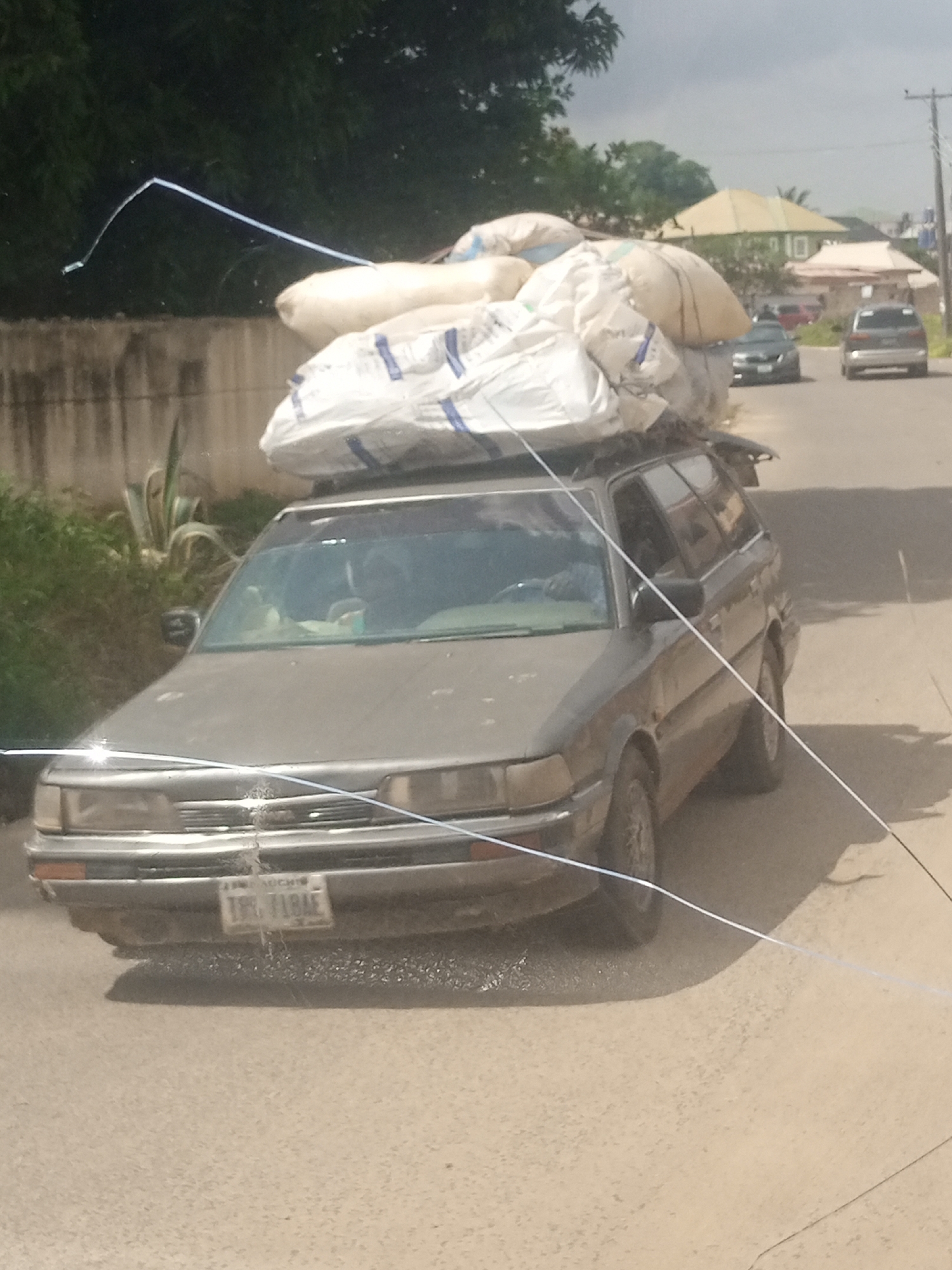 A brown station wagon is loaded with large, white sacks on its roof, secured with straps. The vehicle is driving on a paved road. The sacks appear to be agricultural products or goods. Inside the car, a person is visible in the driver's seat. The scene is set in a location with some vegetation on the left side of the road, including a large, spiky plant. In the background, there are houses with peaked roofs and other vehicles on the road. The sky is overcast, suggesting a cloudy or possibly rainy day. The image is taken from a slightly elevated, side-angle perspective. A license plate is visible on the front of the car, but the text is partially obscured and difficult to read. The overall impression is a scene of transportation and commerce in a rural or semi-urban Nigerian setting.
