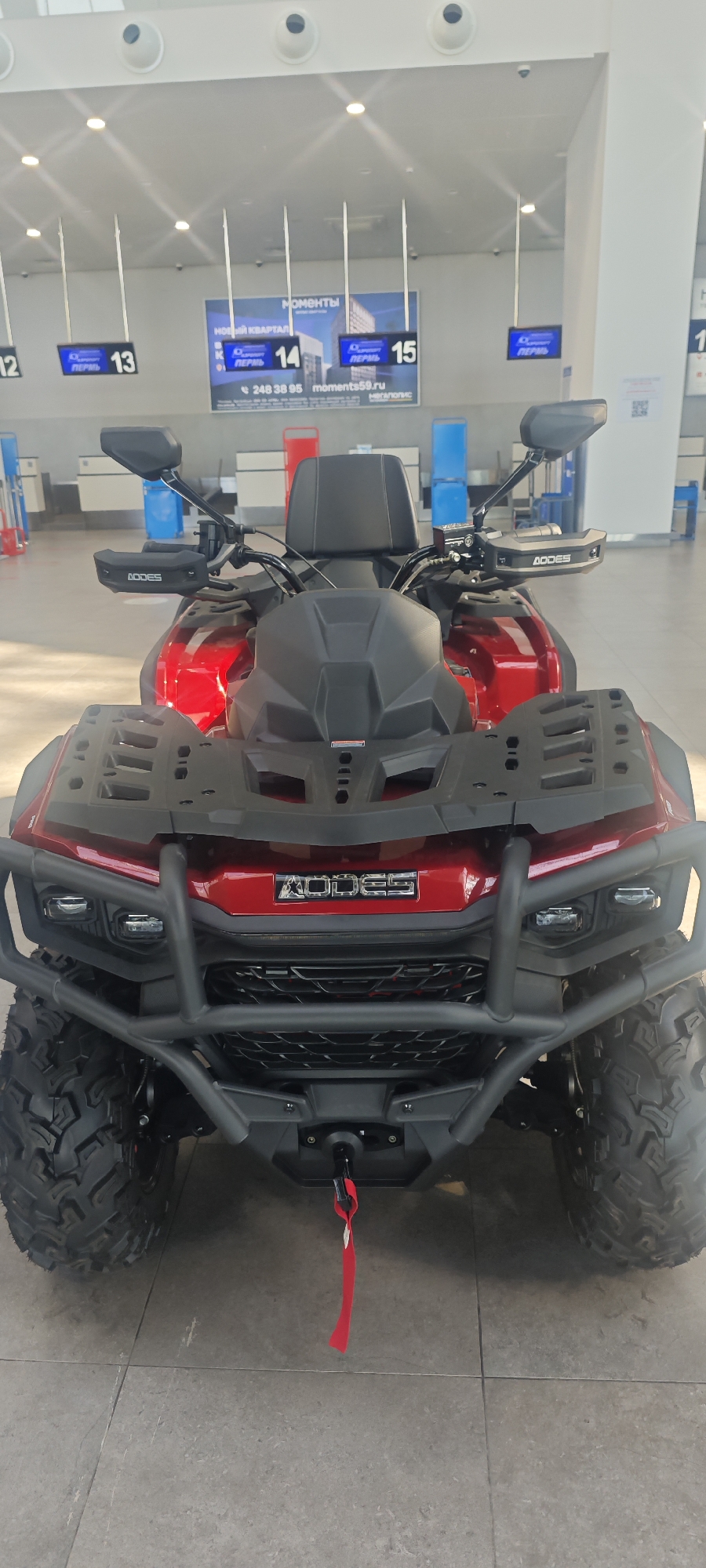 The image shows a red and black AODES brand all-terrain vehicle (ATV) parked indoors. In the background, signs indicate that the ATV is located in Bolshoye Savino Airport, Russia, with signs labeled "Пермь" (Perm) above check-in counters numbered 12 through 15. A phone number and website address are also visible.