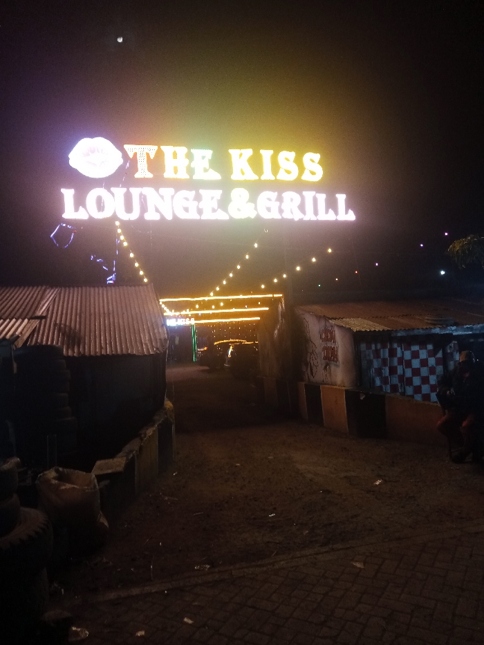 An outdoor view at night featuring a neon sign for "The Kiss Lounge & Grill" with a stylized lip logo. String lights illuminate the entrance area between two structures, one with a corrugated roof and the other checkered. The ground appears uneven, partially paved with bricks, and littered with debris. Tire stacks are visible on the left. The lighting is dim and creates a somewhat gritty atmosphere.