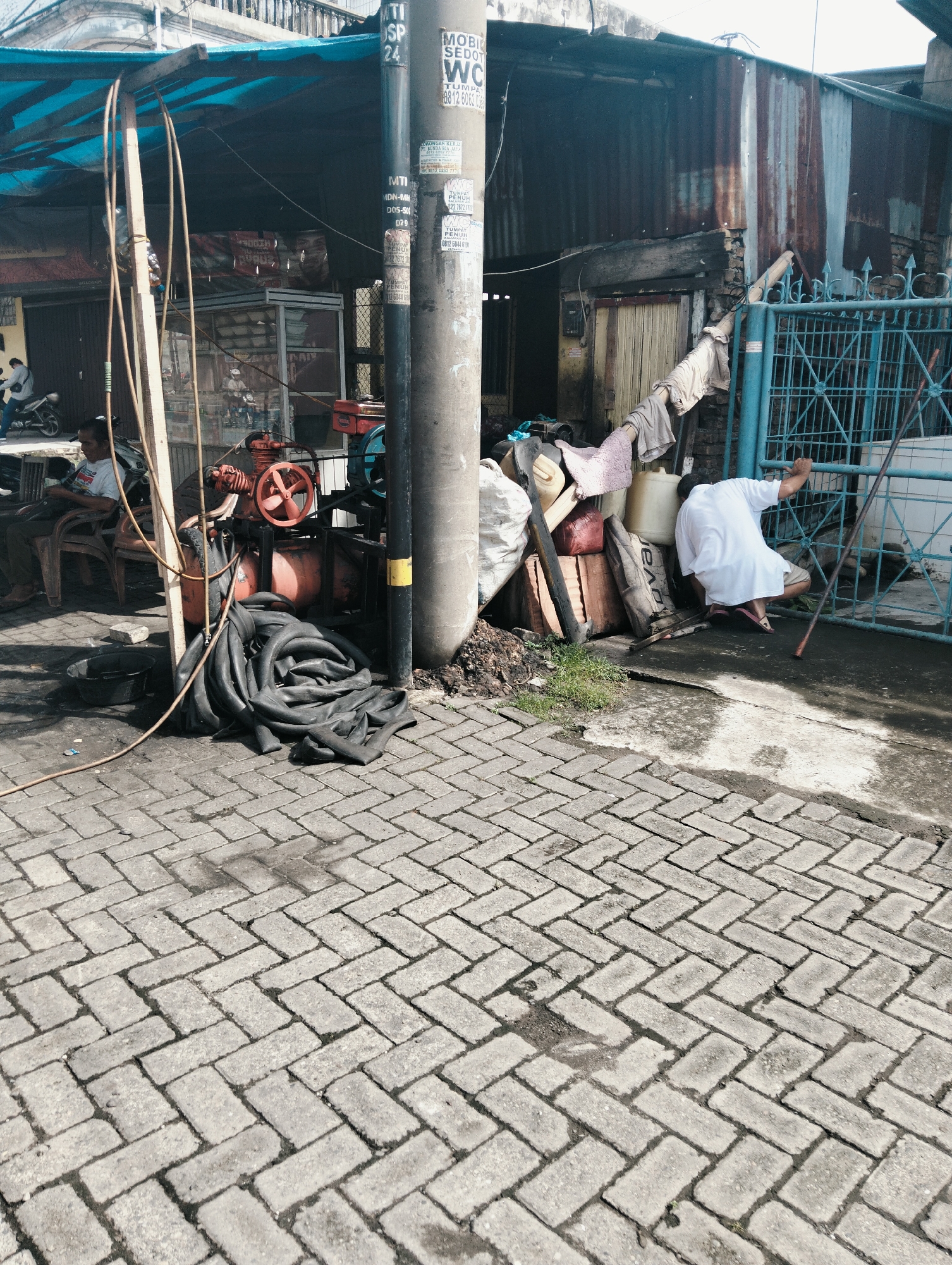 In the City of Medan, Indonesia, a street scene unfolds on a grey, herringbone-patterned brick pavement. To the left, a man sits on a plastic chair under a blue tarp canopy, his torso turned away from the camera and his head tilted slightly down. In front of him, a black plastic basin rests on the ground, and a yellow hose stretches across the pavement towards a pile of black rubber tubes. Adjacent to the tubes is an orange and red air compressor.
Further right, a concrete pole stands vertically, adorned with various stickers. Behind the pole, a rustic building with a corrugated metal roof and walls is visible. A bright blue metal gate stands in front of the building.
A person wearing a white t-shirt and light-colored shorts is crouched near the blue gate, reaching towards it with their right hand. They are wearing sandals. To their left, a long, thin pole rests on the ground. A collection of various items, including plastic containers, sacks, and what appear to be bricks, are stacked against the building wall. The overall atmosphere is one of an outdoor workspace or stall, likely in a busy urban environment.