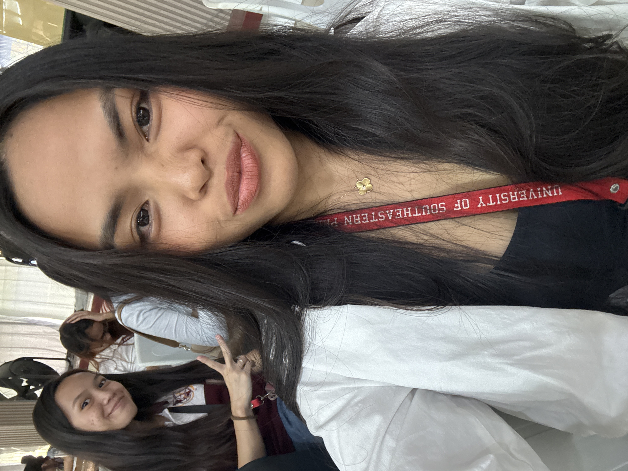 In the foreground, a young woman with long, dark hair takes a selfie. She is wearing a white blazer over a black top and a gold necklace with a flower pendant. A red lanyard with "UNIVERSITY OF SOUTHEASTERN PHILIPPINES" printed on it is visible around her neck. Her makeup includes lipstick and eyeliner. To her left and slightly behind her, another young woman with long dark hair is smiling and making a peace sign with her hand. She is wearing a maroon and white collared shirt. In the background, other individuals are visible, along with chairs and what appears to be a fan. The overall setting suggests an indoor event or gathering. The mention of the "University of Southeastern Philippines" indicates the location is likely within or related to this institution, which has campuses in Davao City, Philippines.