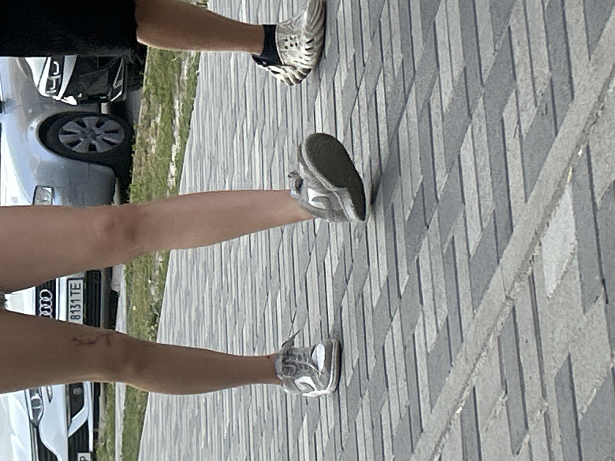 This image captures a low-angle view of two individuals walking on a paved walkway constructed of grey and darker grey rectangular pavers laid in a herringbone pattern. The walkway is flanked by a patch of grass and a street with parked cars. In the background, a white Audi sedan is visible with its license plate reading "8131 TE". To the right of the Audi, a darker colored car, possibly a black SUV, is parked.
The left individual, a person with fair skin and slender legs, is wearing denim shorts and grey Nike sneakers. A faint red mark, potentially a small injury or a temporary tattoo, is visible on their left knee. Their right foot is mid-stride, with the sneaker in focus.
The right individual, whose lower legs are partially visible, is wearing black shorts and distinctive white, open-toed sandals with a textured sole and dark inner lining. This individual's right foot is also visible, placed on the walkway.
The overall scene appears to be an outdoor urban environment, likely in Brovary, Ukraine, given the context provided. The lighting suggests it is daytime.