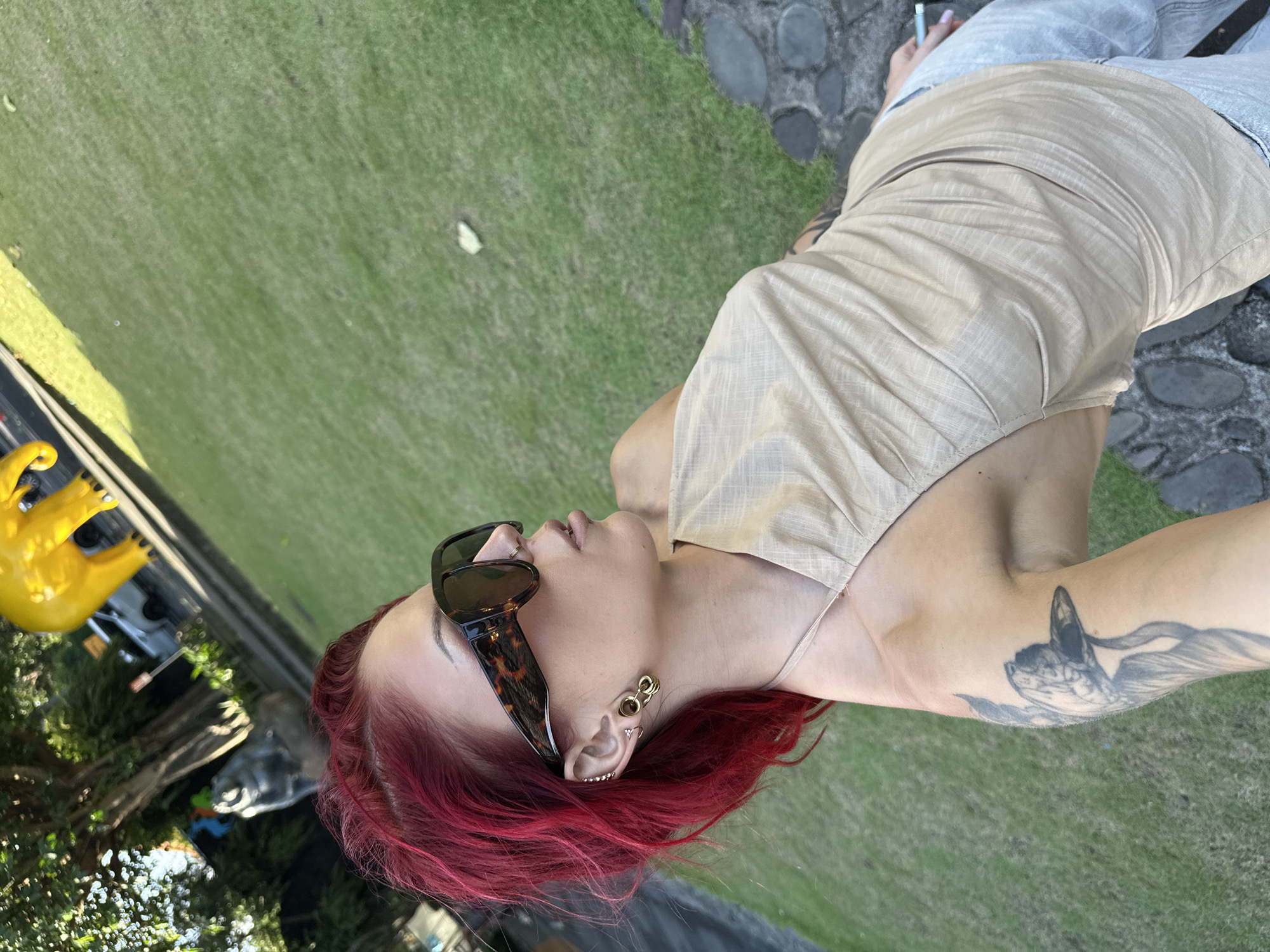 A woman with dyed red hair, wearing sunglasses and a light tan-colored top, poses outdoors. She has multiple ear piercings on her left ear and a tattoo on her left upper arm. She wears light-wash jeans. The location appears to be a grassy area next to a paved walkway, possibly in Sukawati, Indonesia. A yellow decorative item is visible in the background.