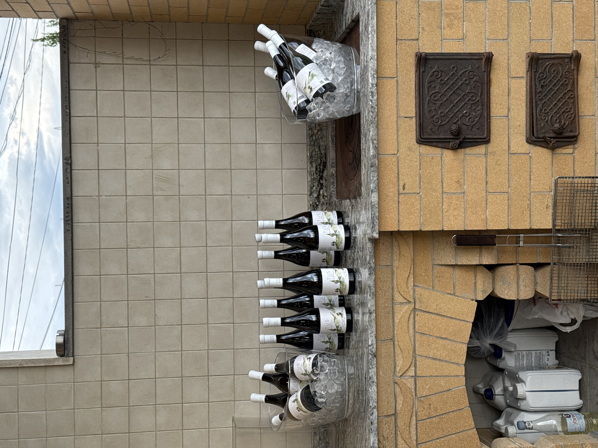 A horizontal medium shot depicts a seemingly exterior section of a structure, with attention drawn to a selection of wine bottles displayed in different manners. Some bottles are nestled in ice-filled containers while others are neatly arranged.
To the right, a rustic brick structure contains a storage nook filled with various household items and equipment. Above this, two decorative, cast-iron panels add a touch of antiquated aesthetic.
To the left, a tiled wall section is visible, framed by what appears to be a window or opening to the outside. The environment is bright, suggesting either daylight or artificial lighting. No people are visible in the frame, and there's no explicit indication of the location being Yerevan, Armenia.