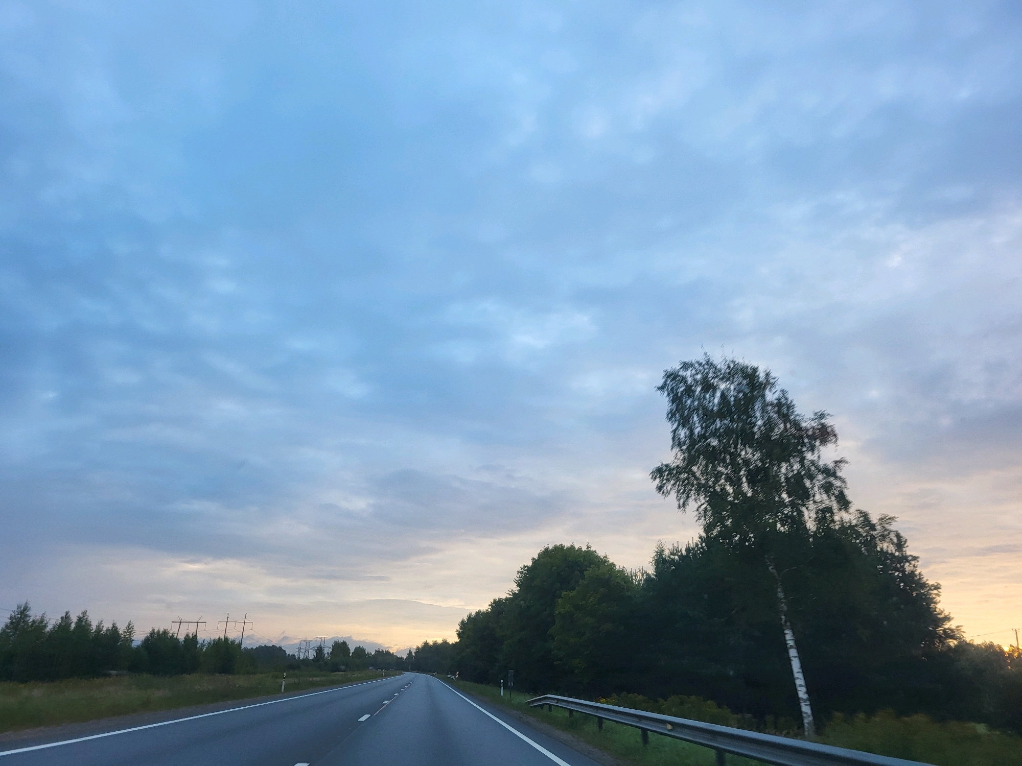 The image depicts a view from a moving vehicle on a two-lane highway in Jukumi, Latvia. The sky is overcast with scattered clouds, showing a gradient of light blue and grey tones, with a hint of yellow and orange on the horizon suggesting either sunrise or sunset.
On the right side of the road, there is a metal guardrail and a dense line of trees, predominantly green with some taller, silhouetted trees. One prominent tree with a white trunk stands out among the foliage, leaning slightly and blurred by motion.
To the left of the highway, there is a grassy shoulder bordered by a line of dark green trees, some of which are shorter and appear to be bushes or young trees. In the distance, beyond these trees, several power lines and utility poles are visible against the sky. The road itself is asphalt and appears to be in good condition, with clear white lane markings. The overall impression is of a rural or semi-rural landscape during twilight hours.