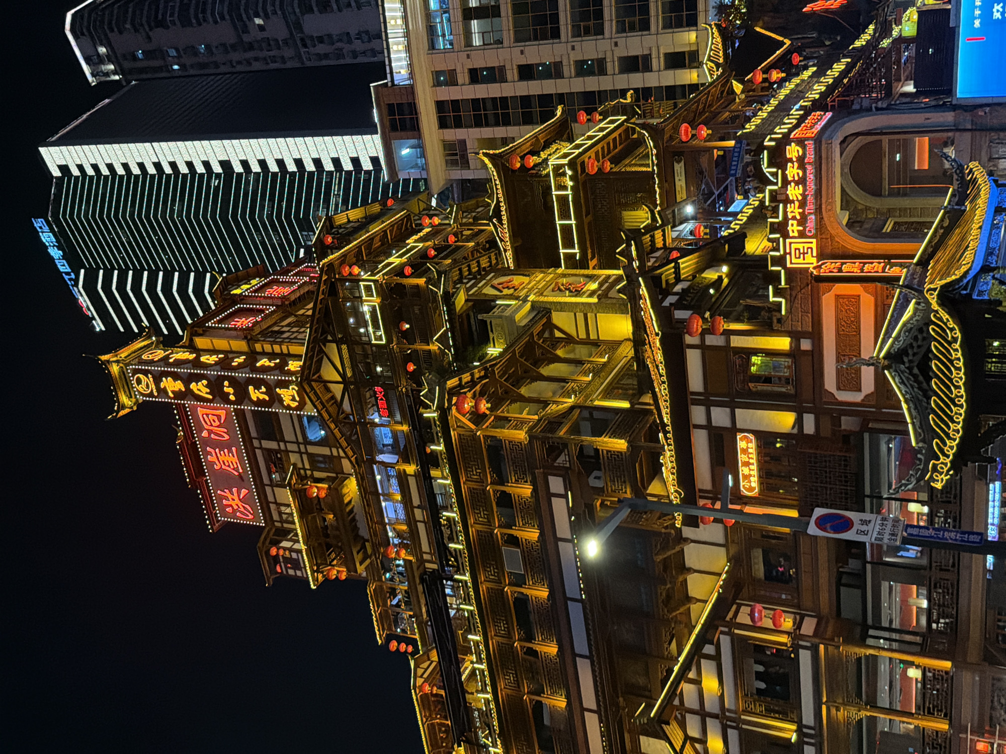 This is a nighttime photograph of a bustling urban scene in the Yuzhong District of China. The primary focus is a multi-story, traditional-style building adorned with an abundance of yellow and red lights, evoking a festive and vibrant atmosphere. The architecture features intricate wooden latticework and several levels of balconies and rooftops, accented by glowing lanterns and illuminated signage.
In the background, a modern skyscraper with a striking geometric pattern of white lights contrasts sharply with the traditional architecture in the foreground. The dark sky suggests the photograph was taken after sunset.
Visible text includes Chinese characters and English, such as "China Time-honored Brand," "洪崖洞" (Hongya Cave), and "江四季酒店" (Jiangsiji Hotel), indicating commercial activity and a specific location within the district. There is also signage related to traffic regulations, including "限时6分钟 (含通行时间)" (Limited to 6 minutes, including travel time) and "禁止停车" (No parking), along with what appears to be directional information. The overall impression is one of a lively, illuminated cityscape where traditional and modern elements coexist. The scene appears busy and perhaps a tourist destination, given the prominent branding and lighting.