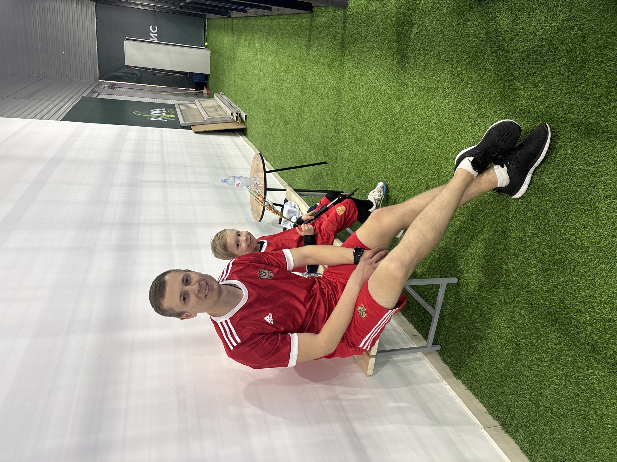 Two people wearing matching red and white Adidas soccer uniforms are posing for a photograph inside what appears to be a sports facility or training center in Chaikovsky, Russia. The older individual, presumably the father, is sitting on a small, wooden stool near a white wall. He is smiling at the camera. His legs are extended, and he is wearing black athletic shoes. A young child, likely his son, is seated on another chair behind him, also dressed in the same uniform. A small table is next to the child, holding a bottle of water and some other objects. The floor is a combination of white flooring and artificial green turf, commonly used in indoor sports facilities. A section of the facility's background features signage with the word "PADEL" visible. The lighting is bright and even, suggesting it is an indoor environment.